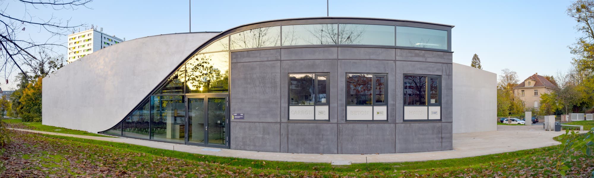 Ein modernes Gebäude mit geschwungener Architektur und großen Glasfenstern. Die Fassade besteht aus grauem Beton und Glas, das die Umgebung reflektiert. Im Vordergrund ist ein gepflegter Rasen mit herbstlichem Laub zu sehen. Im Hintergrund stehen Bäume und ein weiteres Gebäude. Die Szene ist bei Tageslicht aufgenommen.