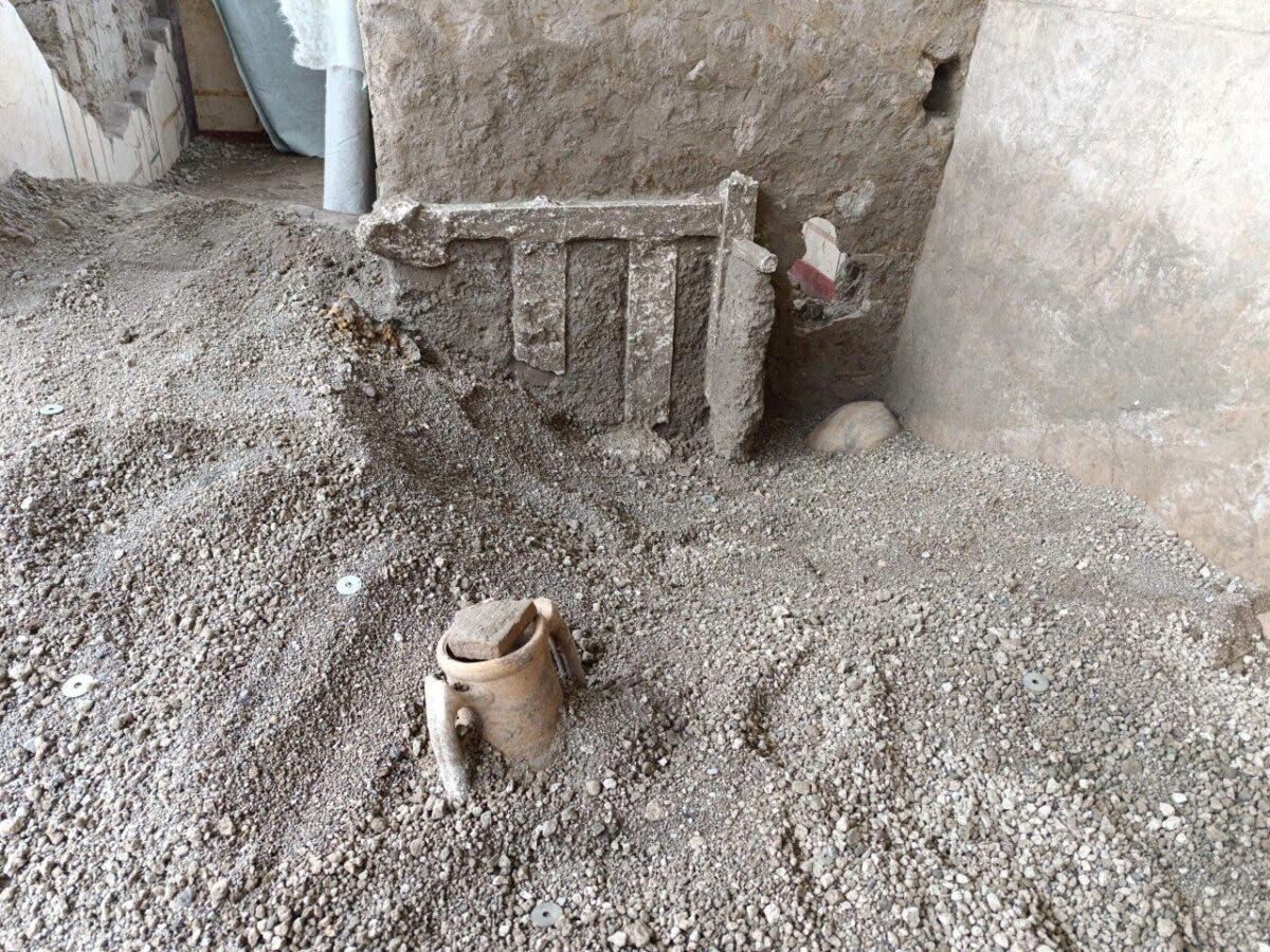 Eine archäologische Ausgrabungsstätte zeigt einen antiken, teilweise frei gelegten Krug, der in einer Schicht aus Kies und Erde eingebettet ist. Im Hintergrund ist eine beschädigte Mauer mit sichtbaren Strukturen zu sehen. Die Szene vermittelt den Eindruck einer laufenden archäologischen Untersuchung.