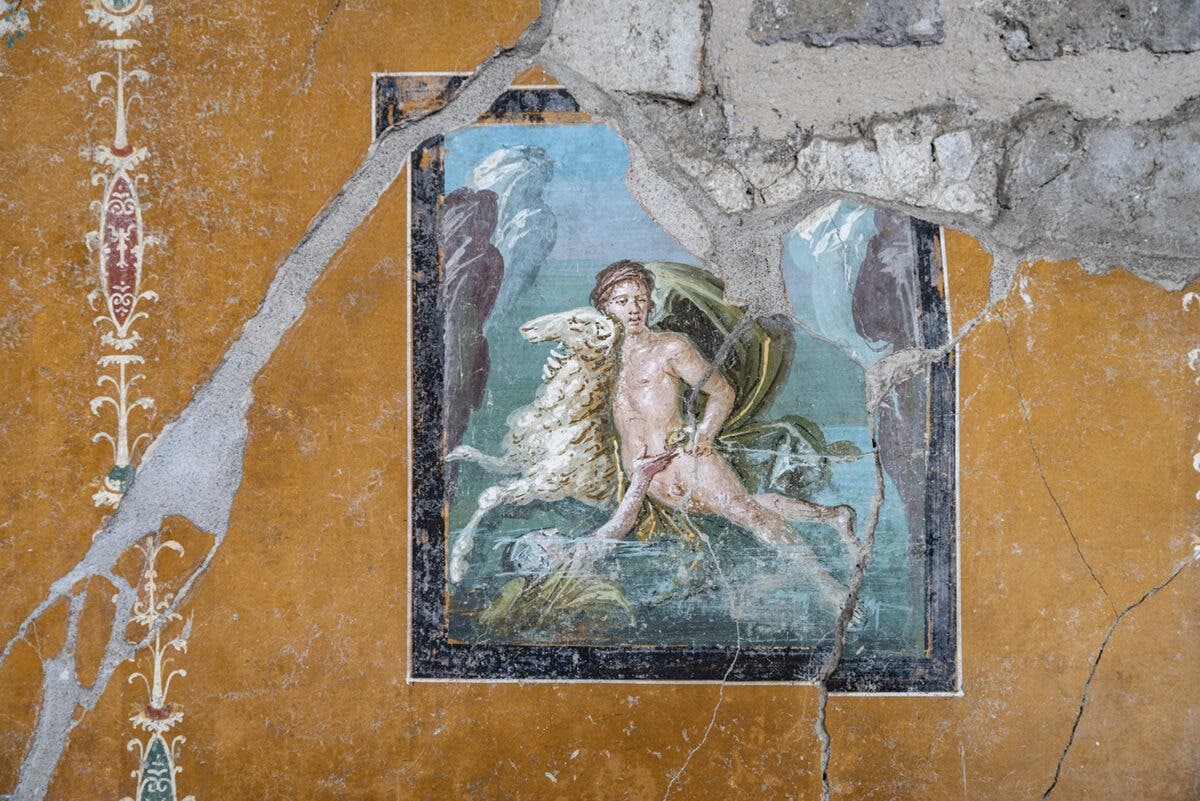 Ein antikes Wandgemälde zeigt eine mythologische Szene mit einer zentralen Figur, die auf einem Widder mit goldenem Fell reitet. Die Figur steht in einer dynamischen Pose vor einem Hintergrund aus Wasser und Felsen. Eine weitere Figur befindet sich unterhalb des Reiters im Wasser und streckt ihre Hände nach ihm aus. Das Gemälde ist von einem orangefarbenen, dekorativen Rahmen umgeben, der Risse und Abnutzungen aufweist. Die Umgebung deutet auf eine historische Stätte hin.