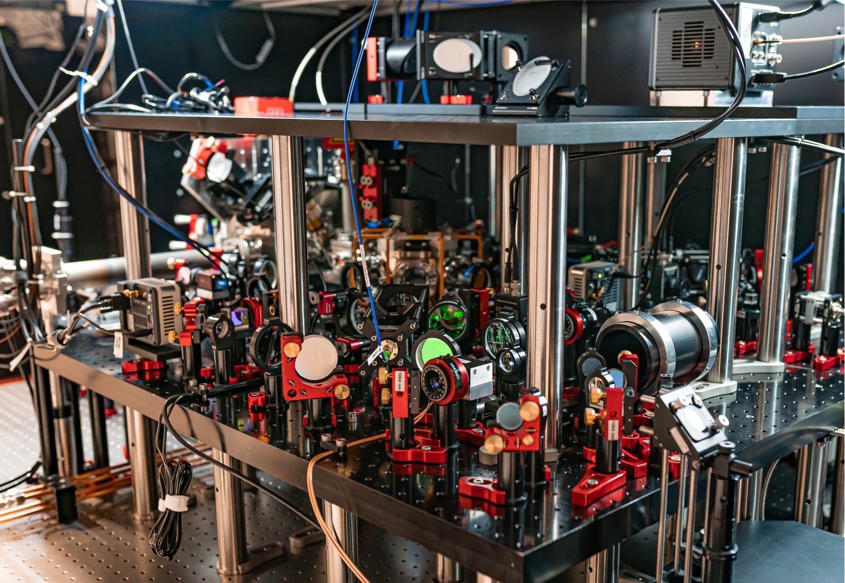 Ein Laboraufbau mit optischen Geräten und Lasern auf einem stabilen Tisch. Verschiedene Linsen, Spiegel und elektronische Komponenten sind auf der Tischoberfläche montiert. Kabel verlaufen über den Tisch, und die Geräte sind in einem komplexen Arrangement angeordnet.