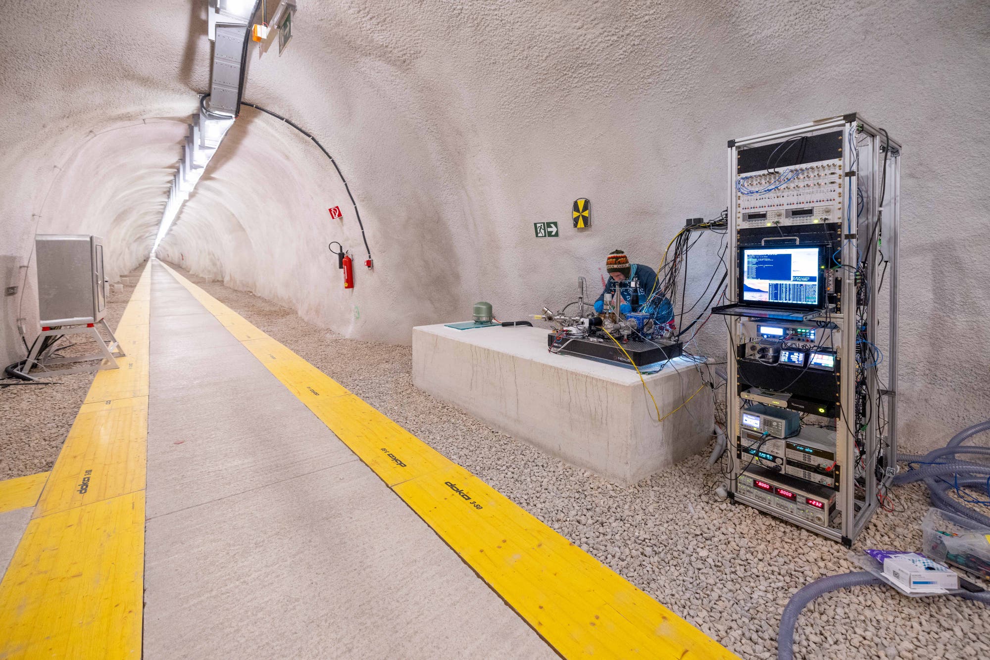Ein Techniker arbeitet in einem langen, beleuchteten Tunnel. Rechts im Bild befindet sich ein großer Metallrahmen mit verschiedenen elektronischen Geräten und Monitoren, die Daten anzeigen. Der Techniker sitzt auf einer Plattform und ist von Kabeln umgeben. Der Boden des Tunnels ist mit gelben Platten markiert, und an der Wand sind Sicherheitszeichen angebracht.