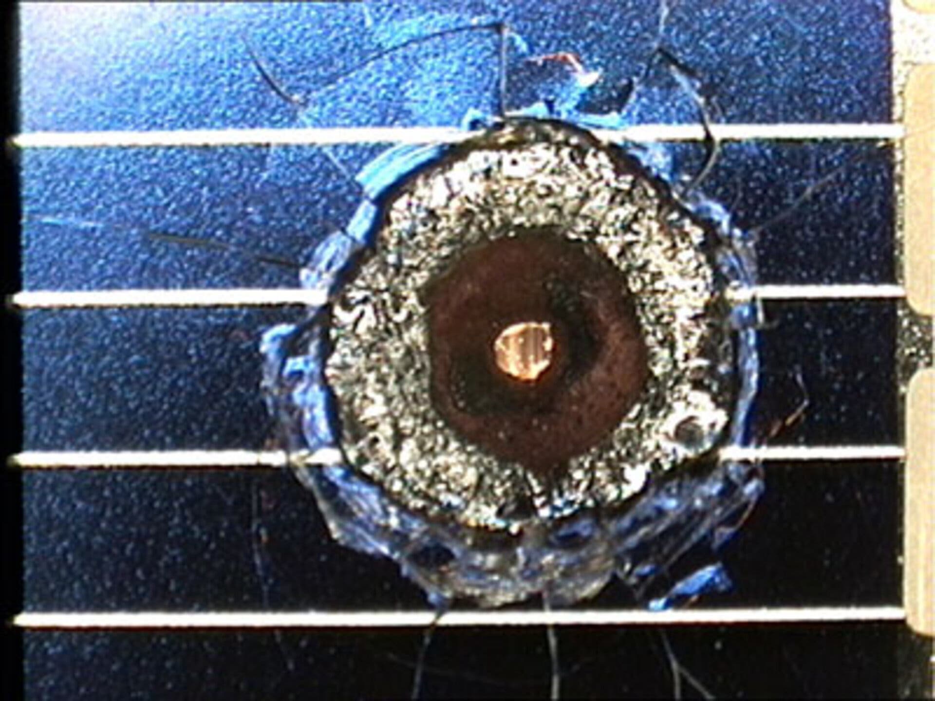 Nahaufnahme eines Mikrometeoriten-Einschlags auf einem blauen Solarpanel. Der Einschlag hat ein kreisförmiges Loch mit verbrannten Rändern hinterlassen. Die Oberfläche des Solarpanels zeigt feine Risse und Kratzer um den Einschlag herum. Das Bild veranschaulicht die potenziellen Schäden durch Weltraummüll an Satelliten.