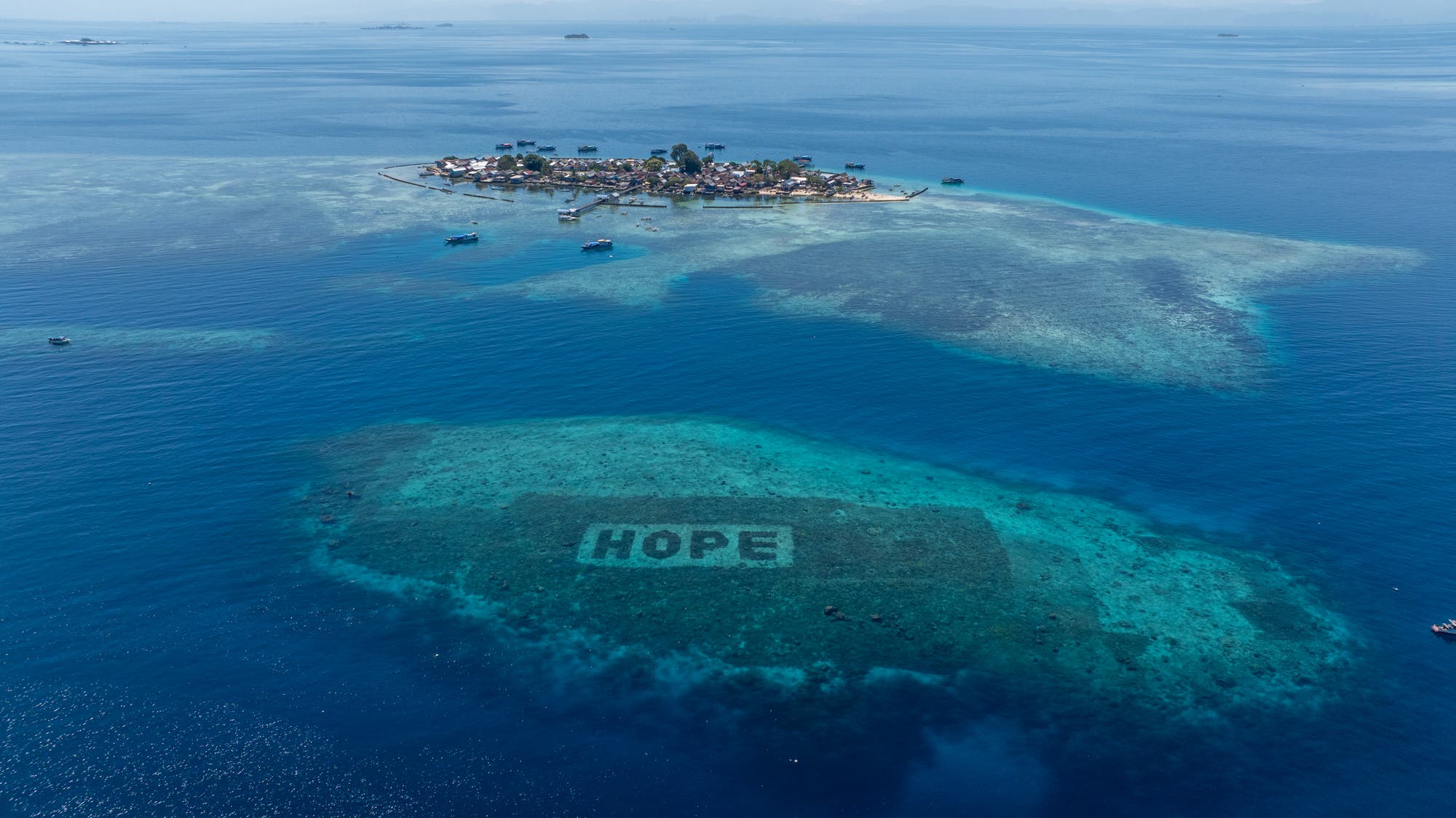 Luftaufnahme des Salisi'-Besar-Riffs in Indonesien. Im Vordergrund ist ein großes, rechteckiges Korallenriff zu sehen, auf dem das Wort »HOPE« in Großbuchstaben sichtbar ist.