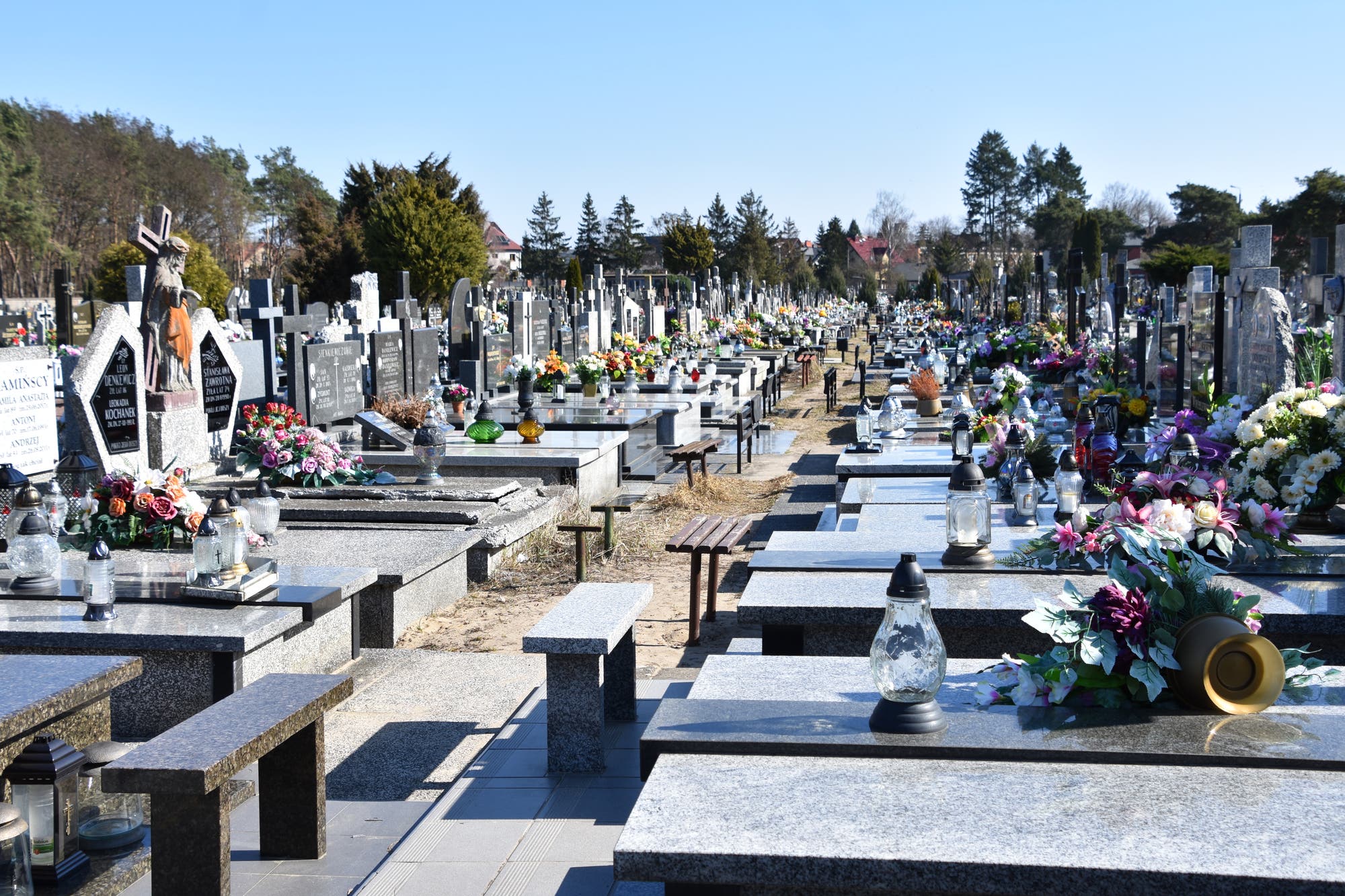 Ein weitläufiger Friedhof mit Reihen von Grabsteinen und Gräbern, die mit bunten Blumenarrangements und Laternen geschmückt sind. Im Hintergrund sind Bäume und einige Gebäude sichtbar. Der Himmel ist klar und blau, was eine ruhige und friedliche Atmosphäre schafft.