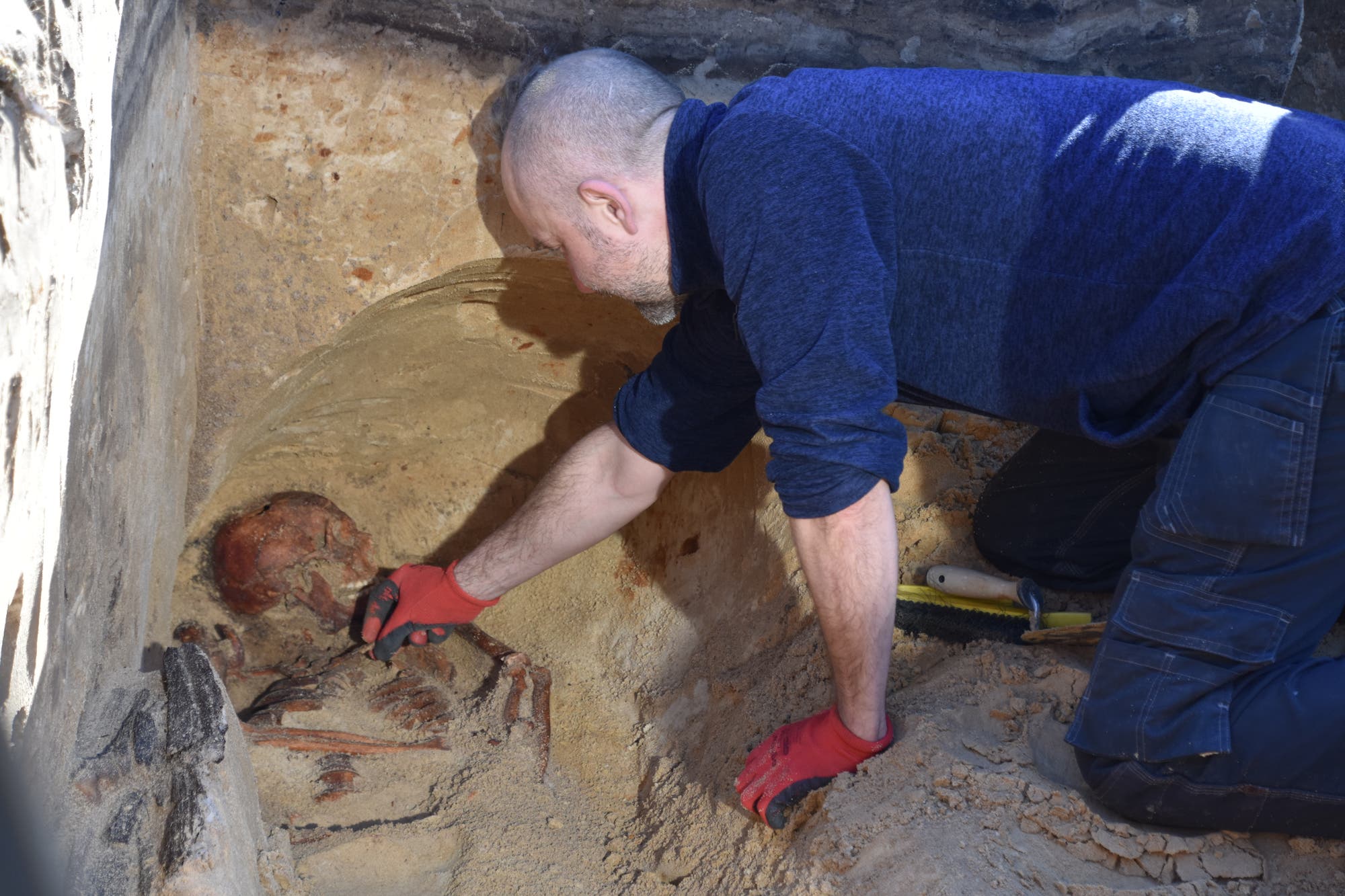 Ein Ausgräber in blauer Kleidung und roten Handschuhen untersucht ein Skelett bei einer Umbettung. Der Mann kniet in einer Grube und verwendet ein kleines Werkzeug, um vorsichtig den Sand um das Skelett zu entfernen. Die Szene ist von einer sandigen Umgebung umgeben.