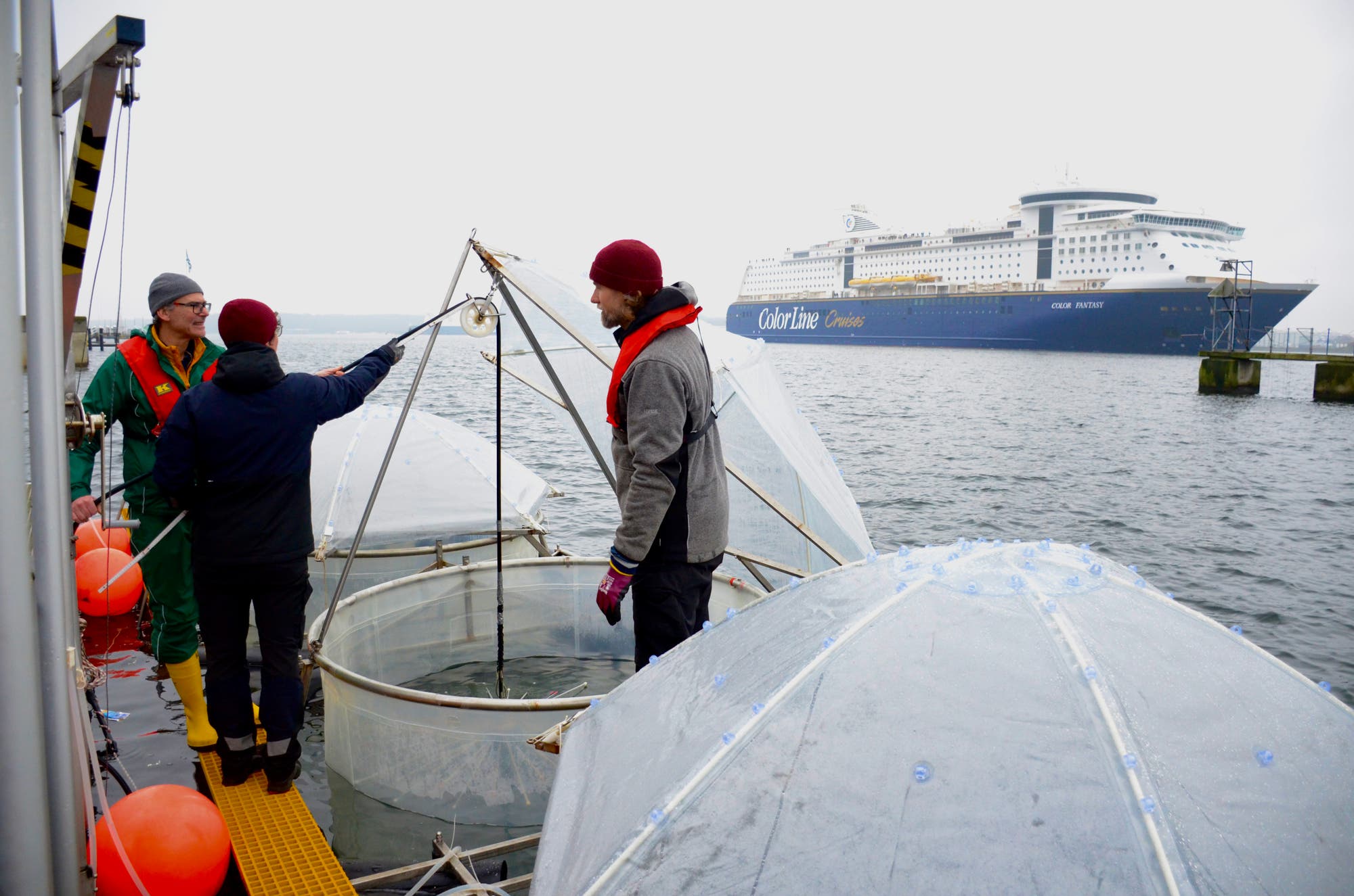 Drei Männer stehen an einem Mesokosmos, im Hintergrund fährt ein Schiff durchs Bild