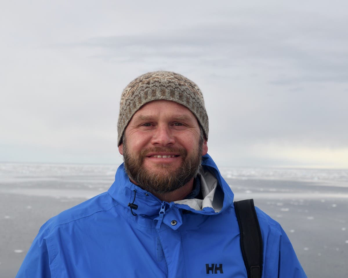 Eine Person in einer blauen Jacke und Strickmütze steht lächelnd vor einer winterlichen Landschaft mit gefrorenem Wasser und bewölktem Himmel.