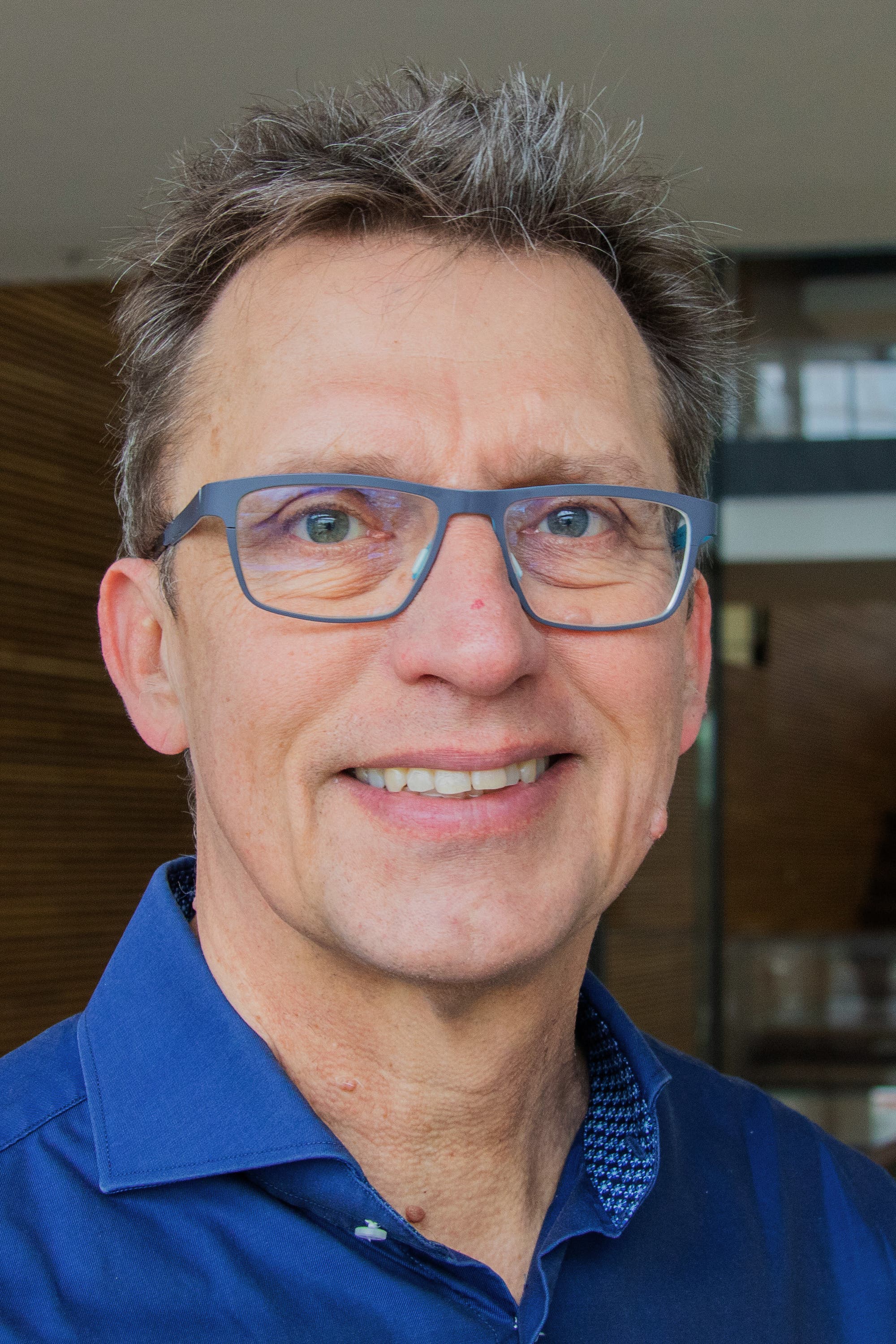 Ein Mann mit Brille und blauem Hemd lächelt in die Kamera. Der Hintergrund ist unscharf und zeigt ein modernes Gebäude.