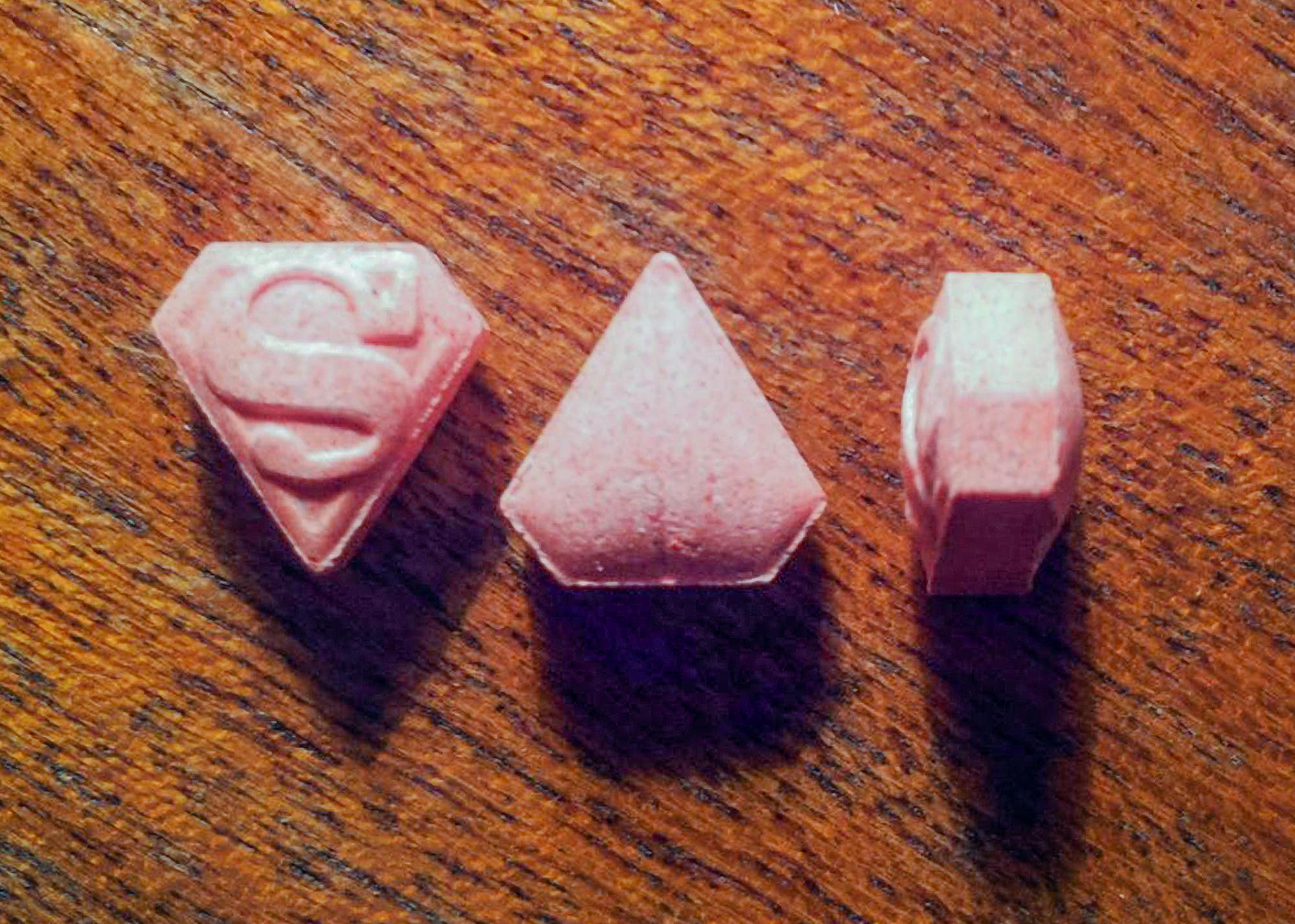 Drei rosafarbene, diamantförmige Tabletten auf einer Holzoberfläche. Die linke Tablette hat ein eingraviertes "S"-Symbol. Die mittlere Tablette ist glatt und ohne Gravur. Die rechte Tablette zeigt eine seitliche Ansicht.