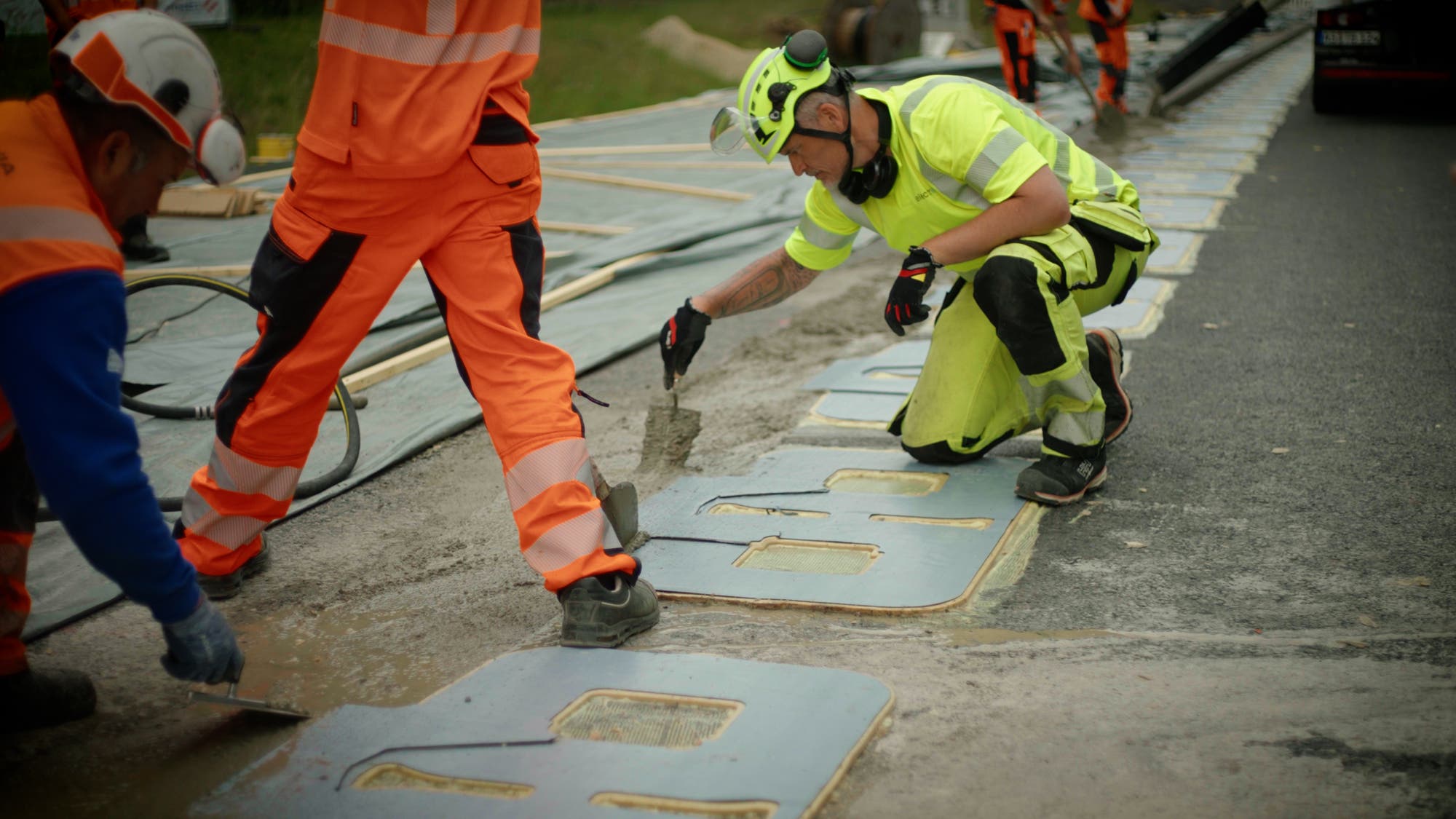 Arbeiter in orangefarbener und gelber Sicherheitskleidung verlegen die flächigen Induktionsspulen hintereinander auf einer Autobahn-Baustelle. Im Hintergrund sind weitere Arbeiter zu sehen. Die Szene zeigt eine Baustelle, auf der Straßenschilder oder Markierungen installiert werden.