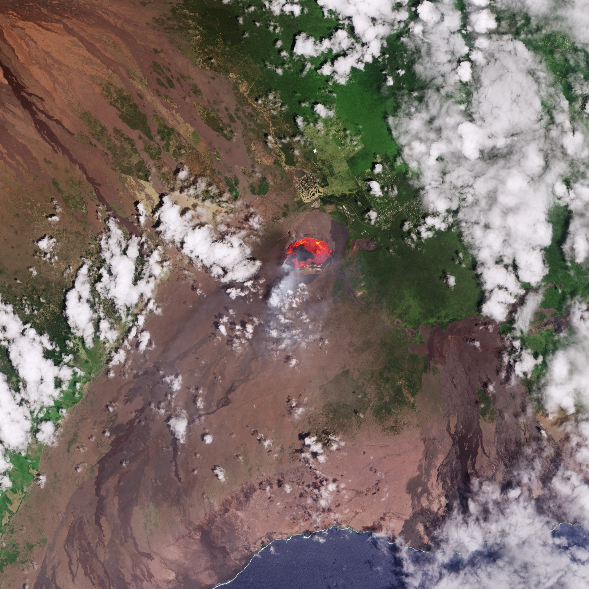 Satellitenbild eines Vulkans mit einem aktiven Lavasee in der Mitte. Die Umgebung zeigt eine Mischung aus gr&uuml;ner Vegetation und braunen, vulkanischen Landschaften. Wei&szlig;e Wolken sind &uuml;ber das Bild verteilt, und die K&uuml;stenlinie ist am unteren Rand sichtbar. Das Bild veranschaulicht die geologische Aktivit&auml;t und die nat&uuml;rliche Umgebung des Vulkans.