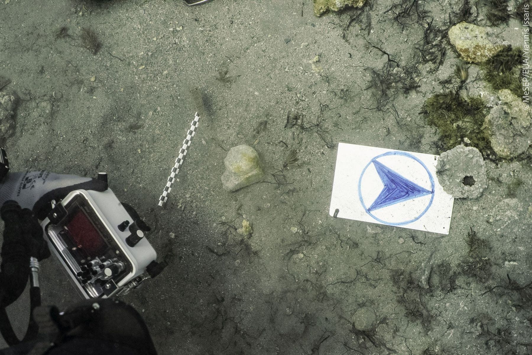 Unterwasseraufnahme eines sandigen Meeresbodens mit einem St&uuml;ck Marmor und einer Kamera im Vordergrund. Auf dem Boden liegt eine wei&szlig;e Tafel mit einem blauen Kreis und einem darin gezeichneten blauen Pfeil. Daneben befindet sich ein Ma&szlig;stab. Um die Tafel herum sind Algen und Steine zu sehen.