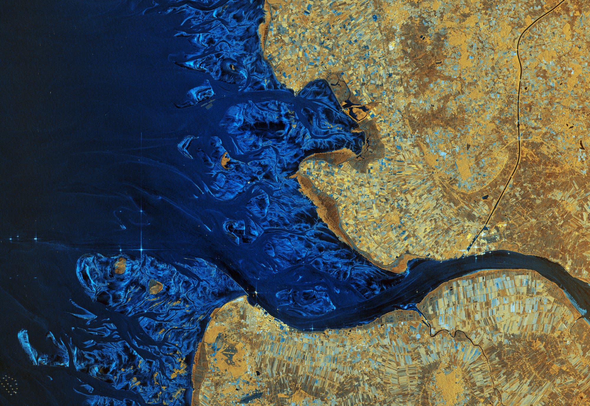 Satellitenbild einer K&uuml;stenregion mit dem Flussdelta der Elbe, die in ein tiefblaues Meer m&uuml;ndet. Die linke Bildh&auml;lfte zeigt das Meer mit verzweigten Wasserwegen, w&auml;hrend die rechte Bildh&auml;lfte landwirtschaftlich genutzte Fl&auml;chen in verschiedenen Braunt&ouml;nen darstellt. Die Karte hebt die Kontraste zwischen Wasser und Land hervor und zeigt die nat&uuml;rliche Geografie der Region.