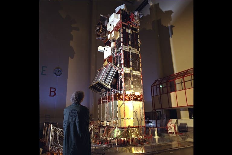 Eine Person in einem Laborkittel steht in einer großen Halle und betrachtet einen hohen Satelliten, der von Kabeln umgeben ist. Die Struktur ist komplex und mit verschiedenen technischen Komponenten ausgestattet. An der Wand im Hintergrund sind die Buchstaben "E" und "B" sowie eine Uhr zu sehen. Die Szene vermittelt den Eindruck von technologischem Fortschritt und Raumfahrtforschung.