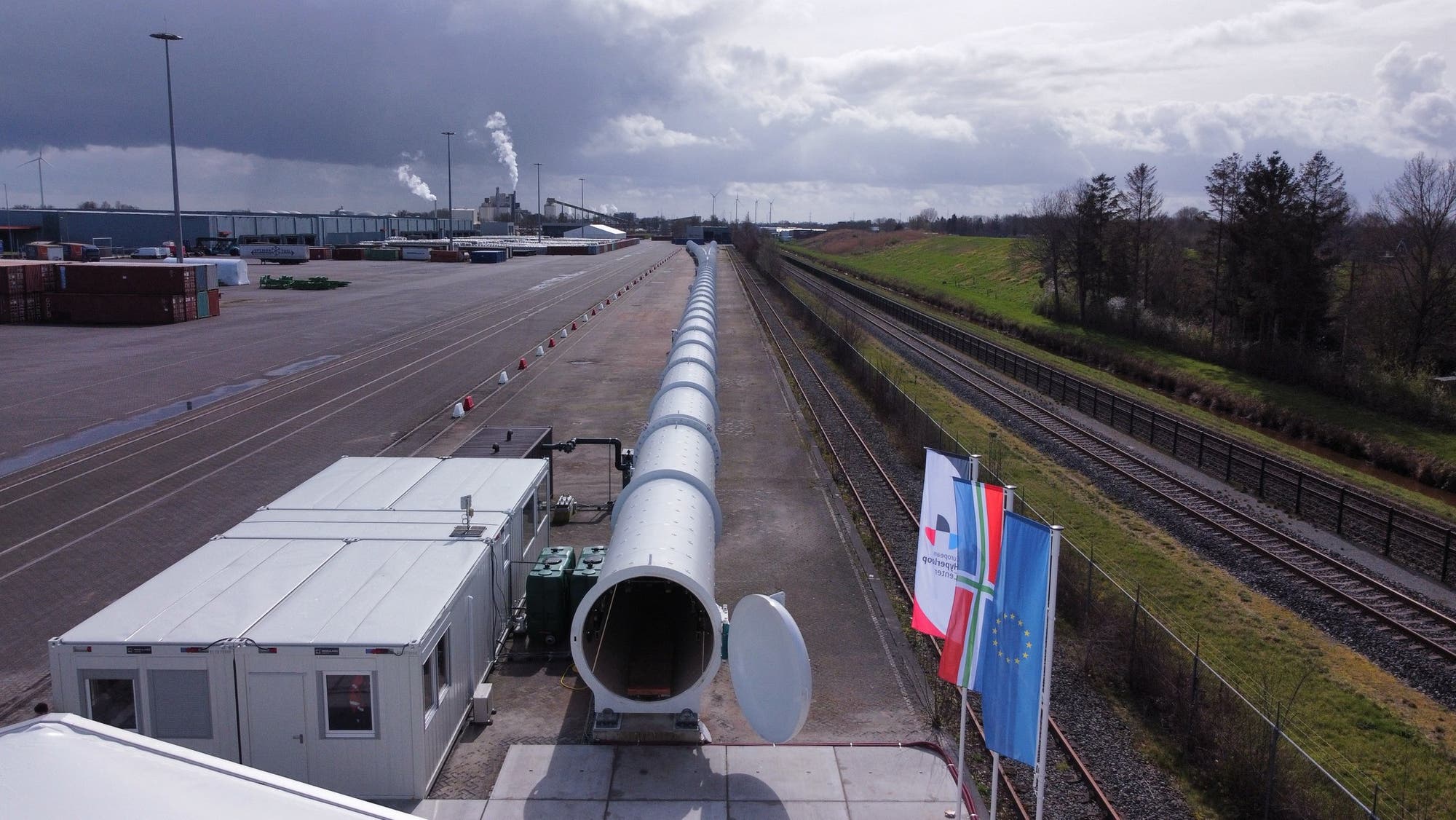 Eine lange, weiße Röhre erstreckt sich entlang eines Industriegebiets neben Bahngleisen. Im Vordergrund befinden sich Container und technische Ausrüstung. Drei Fahnen wehen im Wind, darunter die europäische Flagge. Im Hintergrund sind Fabrikgebäude mit rauchenden Schornsteinen zu sehen. Der Himmel ist bewölkt.