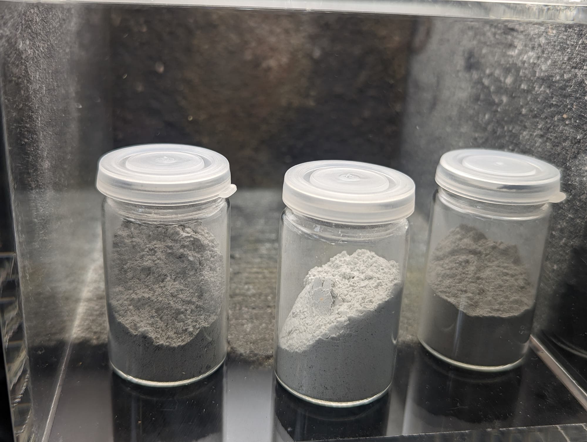 Drei Glasbehälter mit weißem und grauem Pulver, jeweils mit einem weißen Plastikdeckel verschlossen, stehen nebeneinander in einem Regal. Die Behälter enthalten unterschiedliche Mengen an Pulver, das möglicherweise für wissenschaftliche oder industrielle Zwecke verwendet wird. Der Hintergrund ist dunkel und unscharf.