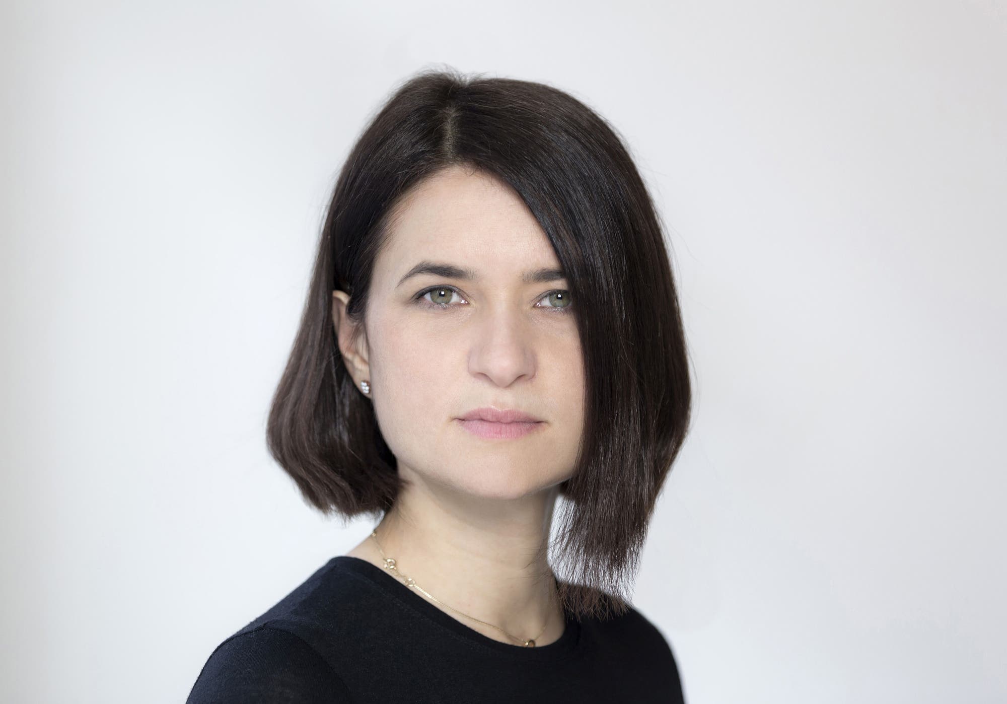 Eine Person mit schulterlangem, dunklem Haar blickt direkt in die Kamera. Sie trägt ein schwarzes Oberteil und hat einen neutralen Gesichtsausdruck. Der Hintergrund ist einfarbig und hell, was den Fokus auf das Gesicht der Person lenkt.