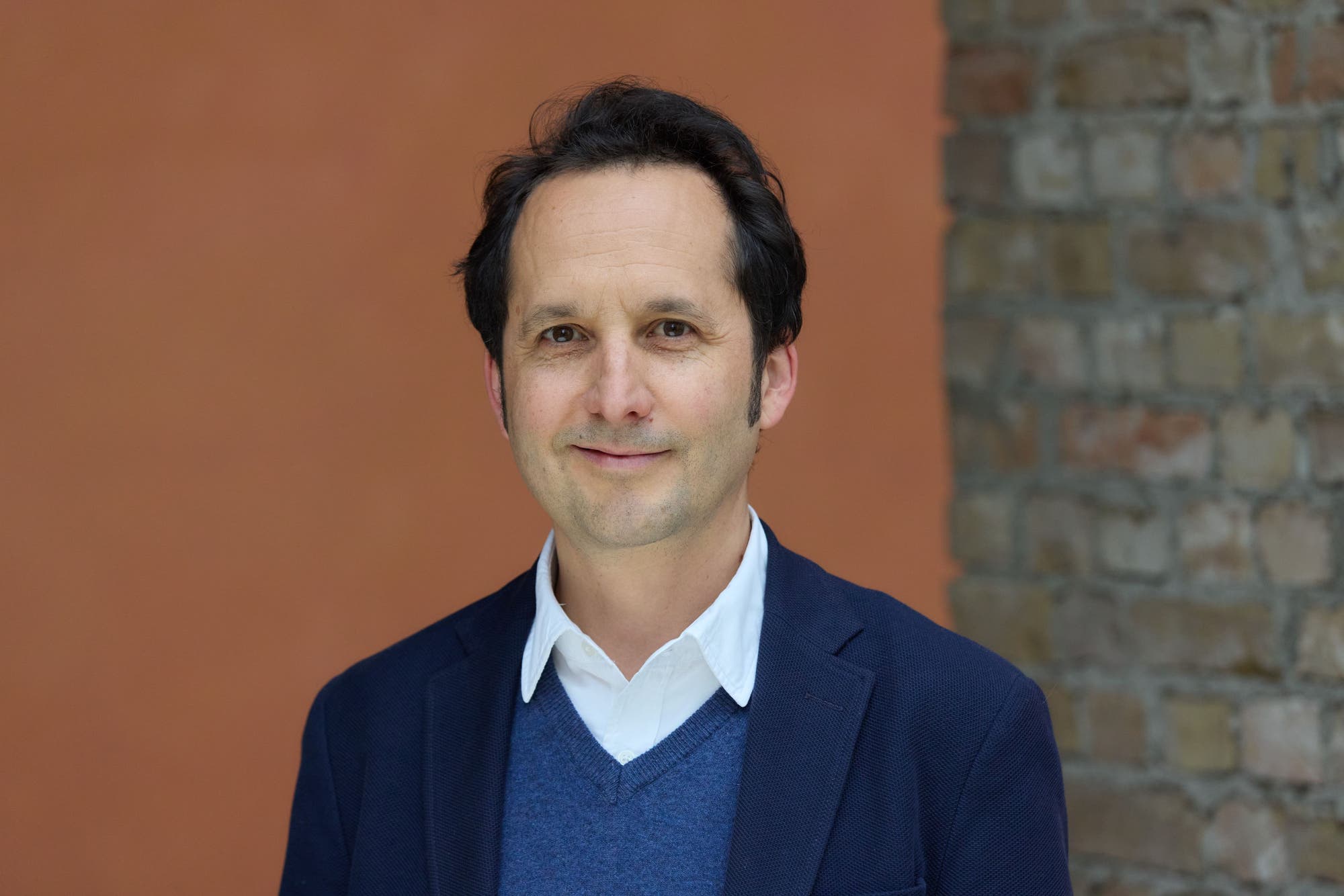 Eine Person in einem blauen Jackett und einem weißen Hemd steht vor einer orangefarbenen Wand und einer Ziegelmauer. Die Person lächelt leicht in die Kamera.