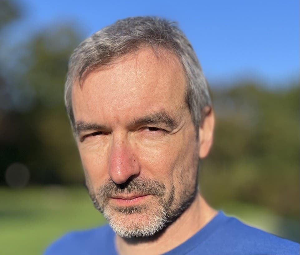 Ein Mann mit grauem Haar und Bart schaut ernst in die Kamera. Er trägt ein blaues T-Shirt. Der Hintergrund ist unscharf und zeigt einen sonnigen Tag mit blauem Himmel und grüner Vegetation.