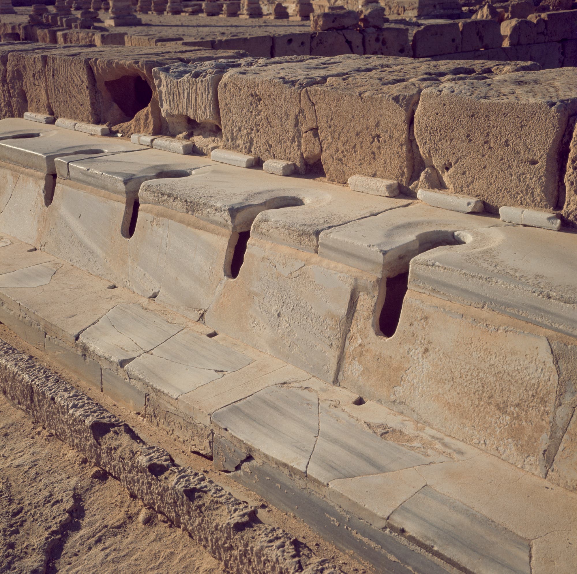 Reihe antiker römischer Toiletten aus Stein, die in einer archäologischen Stätte angeordnet sind. Die Sitze sind aus Marmor gefertigt und zeigen eine gleichmäßige Anordnung mit abgerundeten Öffnungen. Die Struktur ist von verwittertem Gestein umgeben, das die historische Nutzung und den architektonischen Stil der damaligen Zeit widerspiegelt.