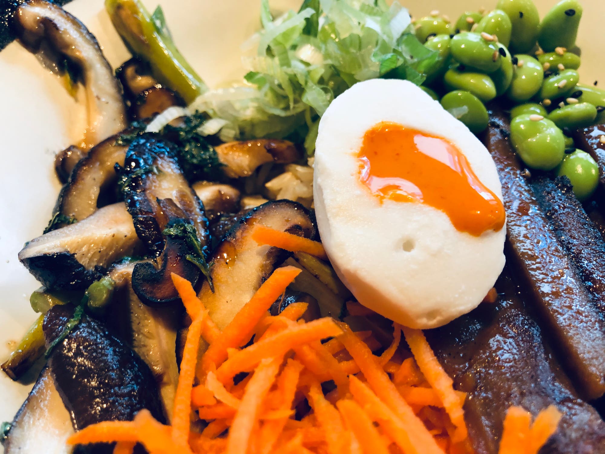 Eine Nahaufnahme zeigt das vegane Imitat eines halben pochierten Eis auf einer Mischung von sautierten Shiitake-Pilzen, Edamame-Bohnen und geraspelten Karotten. Das vermeintliche Eigelb ist orange gefärbt und läuft leicht aus. 