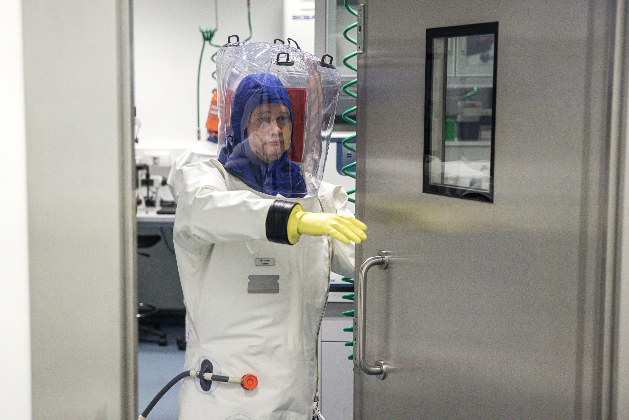 Eine Person in einem weißen Schutzanzug mit transparentem Helm und gelben Handschuhen öffnet eine schwere Metalltür in einem Labor. Im Hintergrund sind Laborgeräte und Schläuche zu sehen. Die Szene vermittelt ein Gefühl von Vorsicht in einer wissenschaftlichen Umgebung.