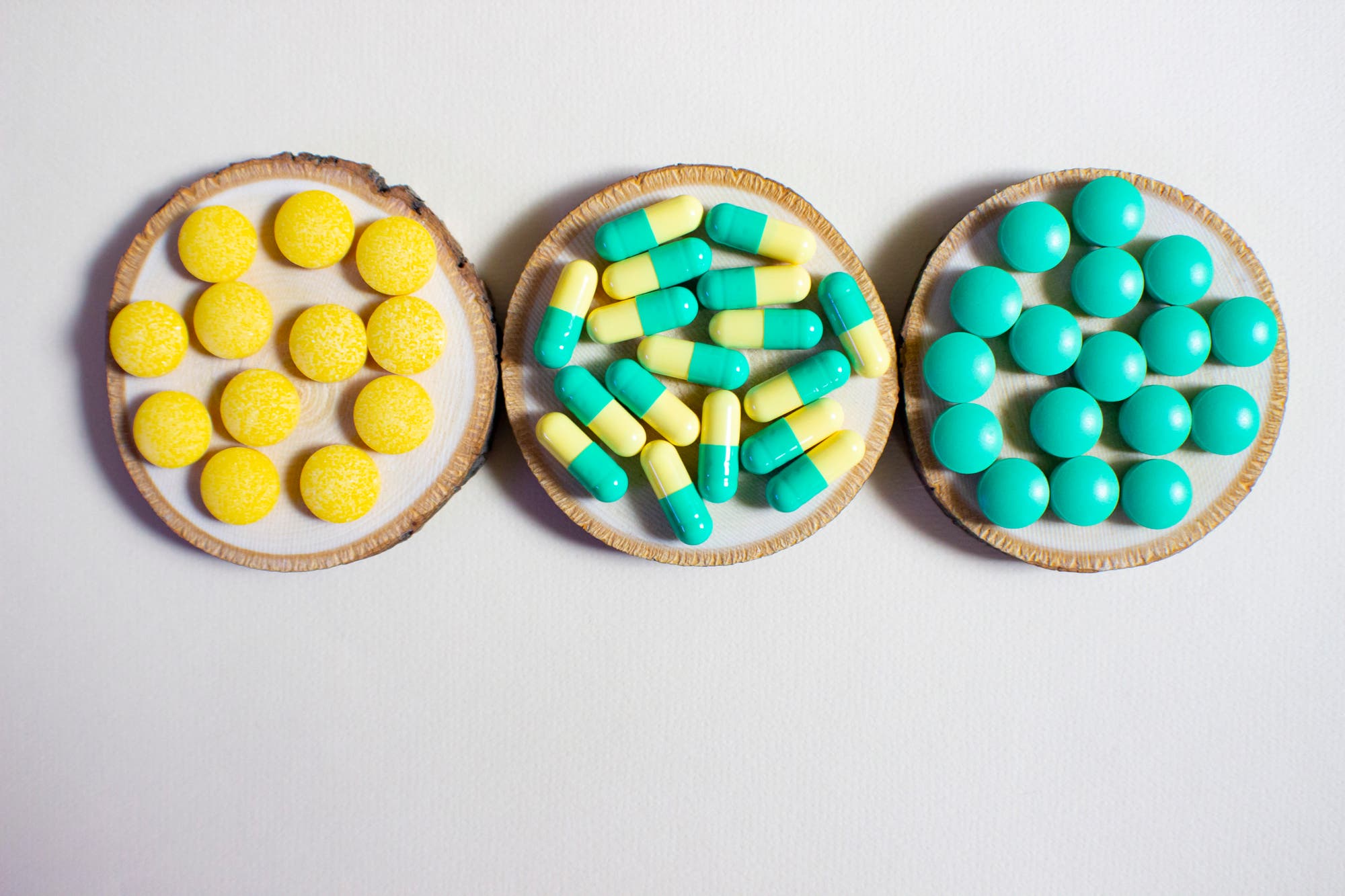 Drei runde Holzuntersetzer auf einer weißen Oberfläche, jeder mit einer anderen Art von Medikamenten: links gelbe Tabletten, in der Mitte gelb-türkise Kapseln, rechts türkisfarbene Tabletten.