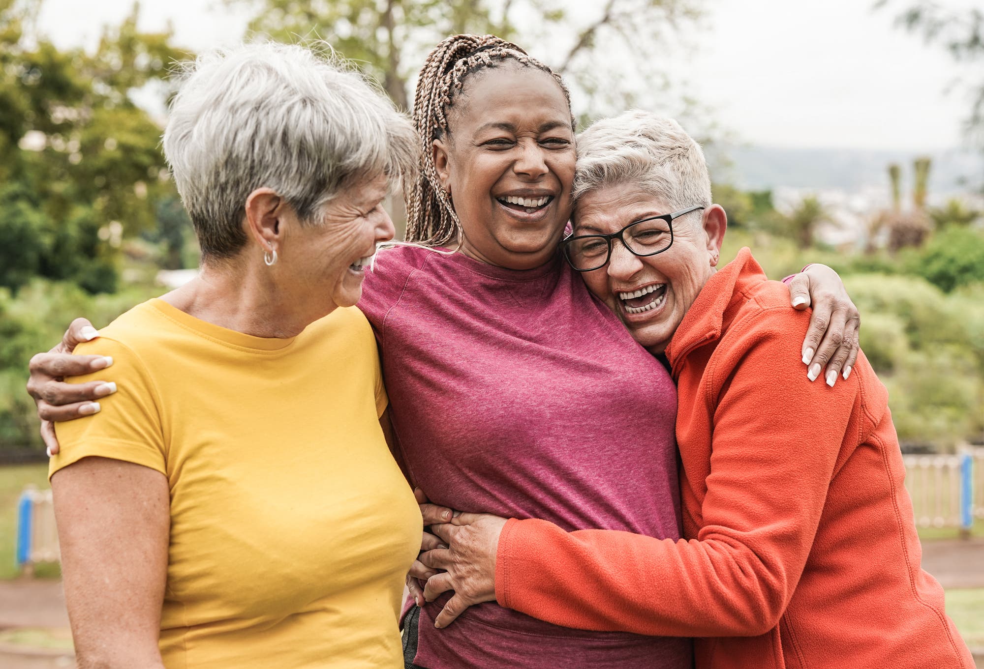 Drei fröhliche Seniorinnen verschiedener Ethnien nebeneinander, die gemeinsam Spaß im Freien haben. Sie lachen und die Frau rechts umarmt diejenige, die sich in der Mitte befindet. Diese wiederum legt die Arme um die beiden anderen.