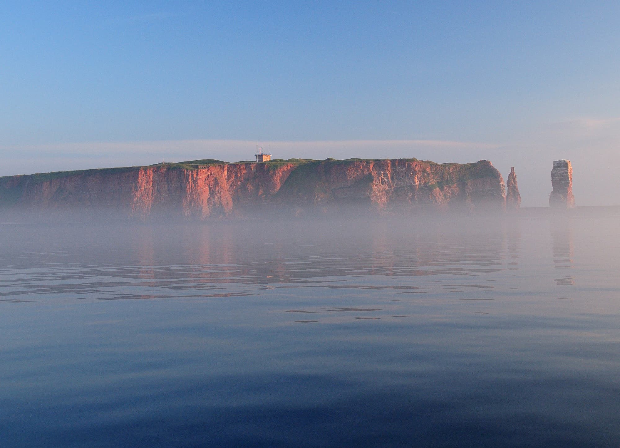 Die felsige Insel Helgoland erhebt sich aus dem ruhigen Meer, umgeben von leichtem Nebel. Die Klippen sind in warmes Licht getaucht. Auf der Insel ist ein Leuchtturm zu sehen. Der Himmel ist klar mit einem sanften Blau.