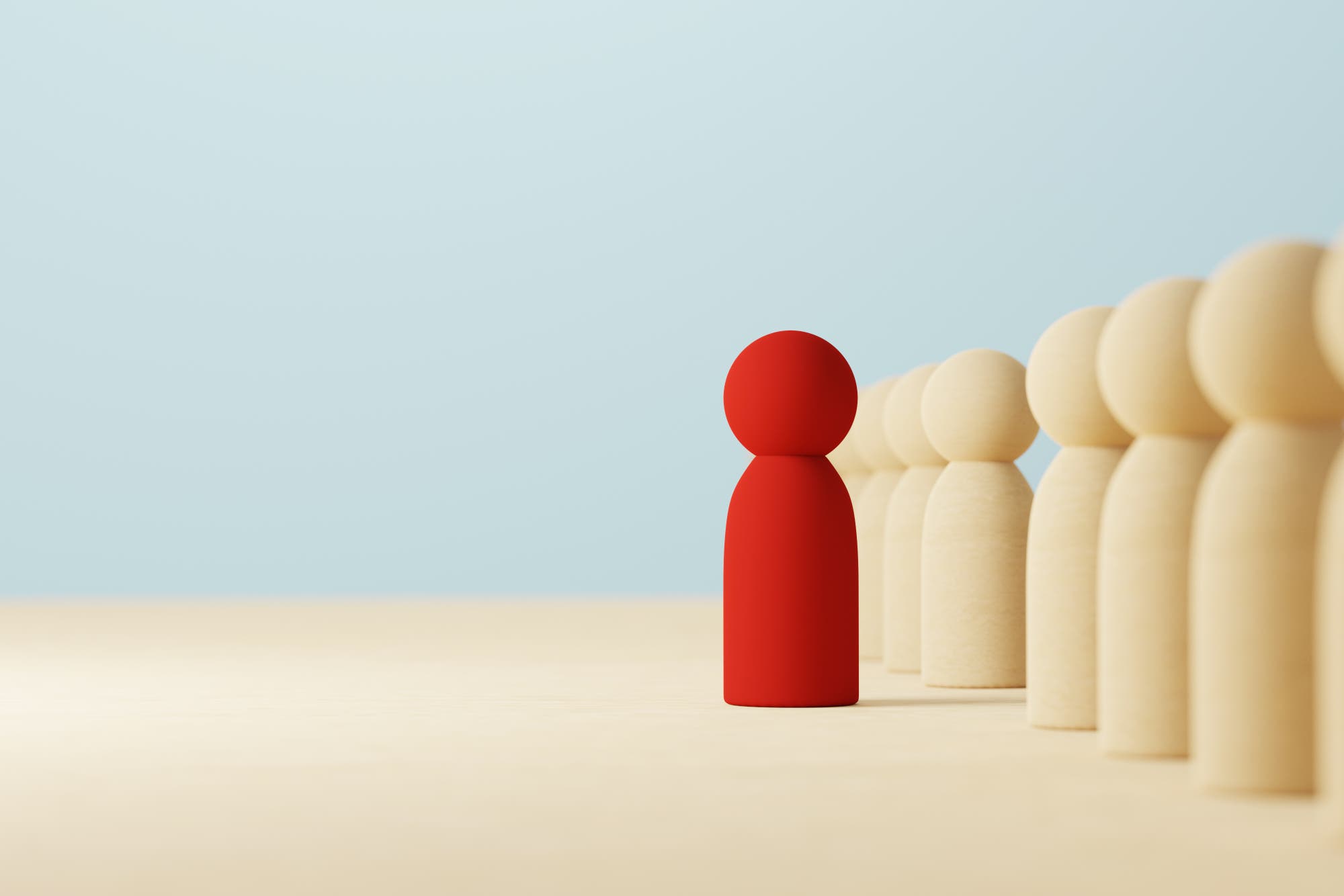 Eine rote Spielfigur steht in einer Reihe von beigen Spielfiguren, die alle in die gleiche Richtung blicken. Die rote Figur hebt sich deutlich von den anderen ab, was Individualität oder Anderssein symbolisieren könnte. Der Hintergrund ist hellblau und schlicht.