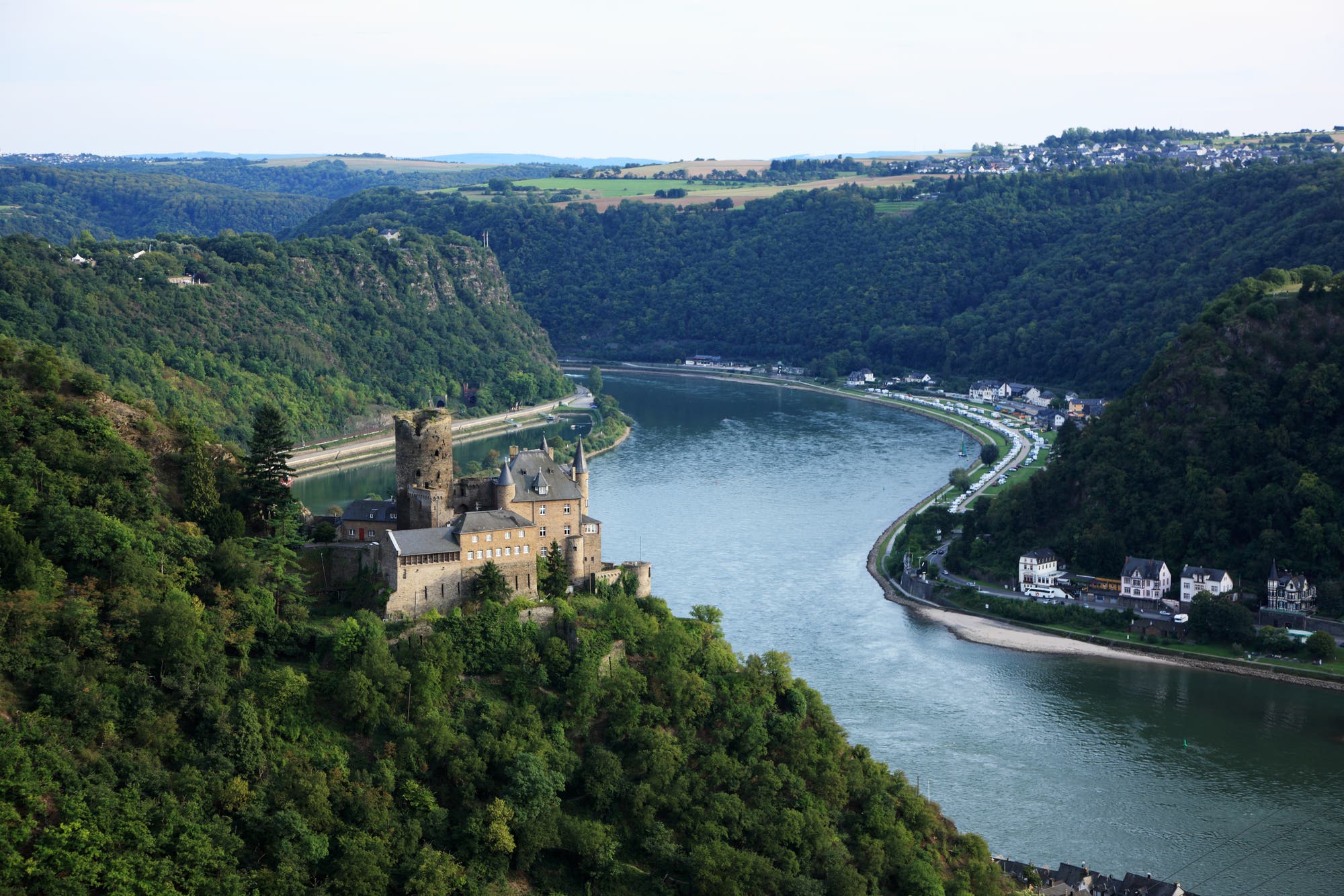 Eine malerische Landschaft mit einer historischen Burg auf einem bewaldeten Hügel, die auf den Rhein blickt. Der Fluss schlängelt sich durch das Tal, umgeben von üppigen grünen Hügeln und kleinen Dörfern am Ufer. Im Hintergrund sind weite Felder und eine Ansammlung von Häusern zu sehen. 