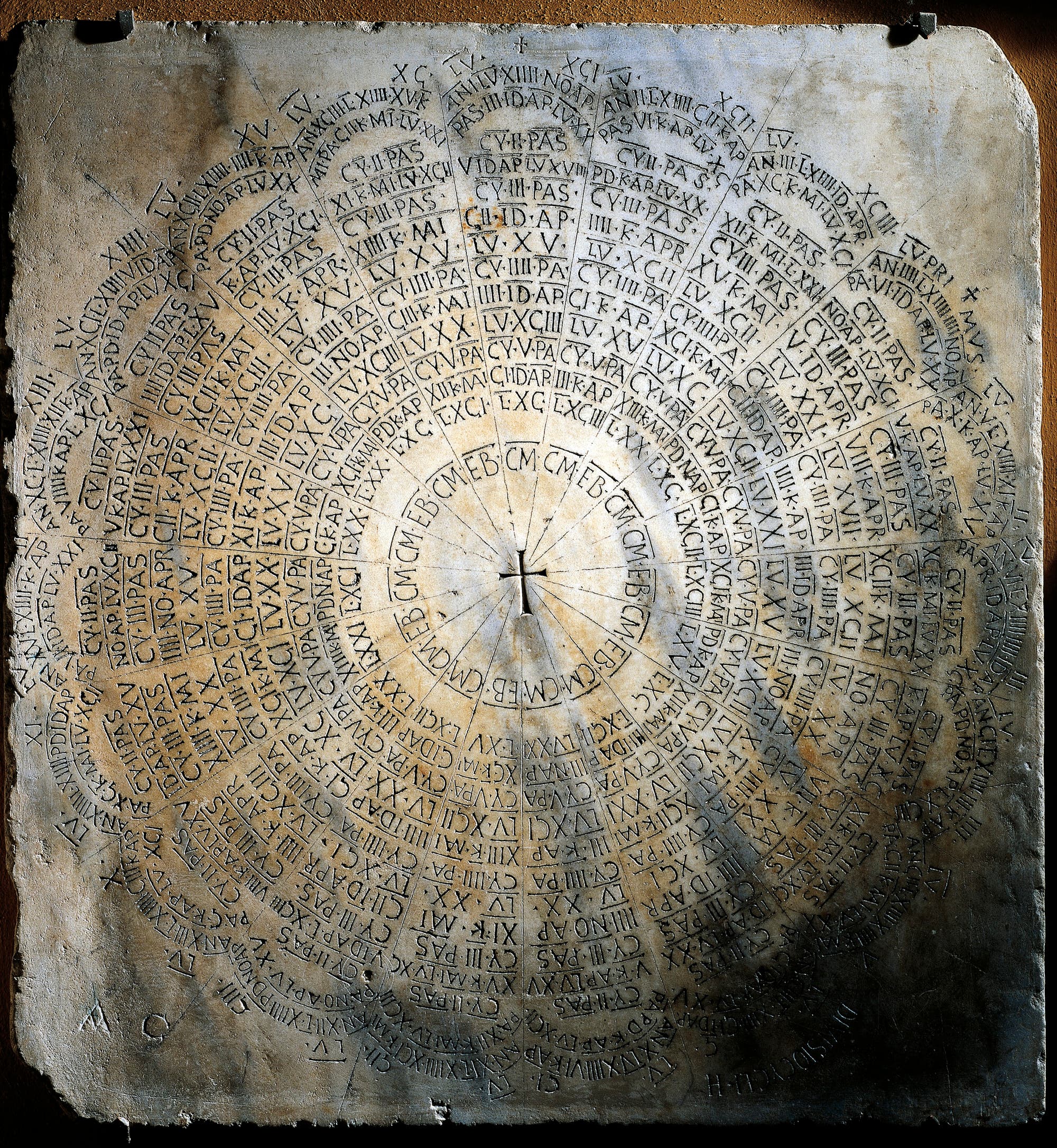 Steinplatte mit kreisförmiger Anordnung von lateinischen Inschriften und römischen Zahlen. Die Inschriften sind konzentrisch um einen zentralen Punkt angeordnet und scheinen eine Art Kalender oder Zählwerk darzustellen. Die Oberfläche ist gealtert und weist Verfärbungen auf.