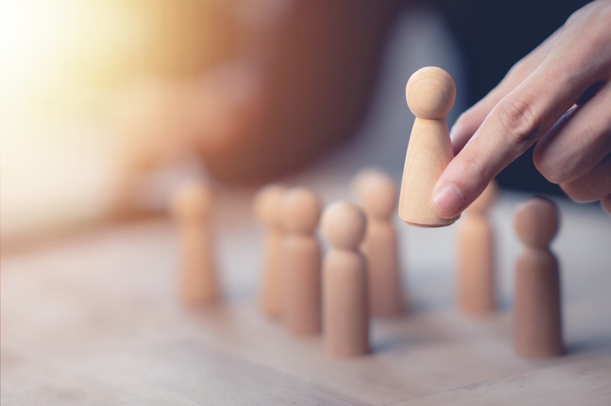 Eine Hand hebt eine hölzerne Spielfigur von einem Tisch, auf dem mehrere ähnliche Figuren stehen. Die Szene ist in warmem Licht gehalten und symbolisiert Führung oder Auswahl.