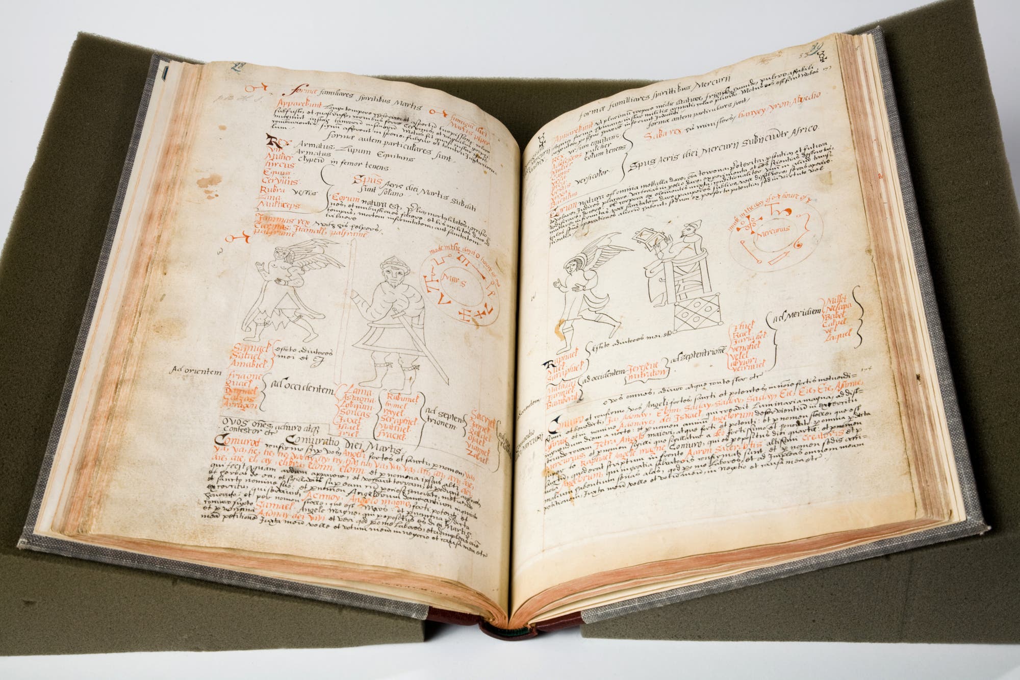 Ein aufgeschlagenes mittelalterliches Manuskript liegt auf einem gepolsterten Buchhalter. Die Seiten sind mit handgeschriebenem lateinischem Text und detaillierten Illustrationen von Figuren verziert. Die Zeichnungen zeigen Personen in mittelalterlicher Kleidung, begleitet von dekorativen Elementen und roten Hervorhebungen. Die Seiten sind leicht vergilbt, was auf das Alter des Dokuments hinweist.