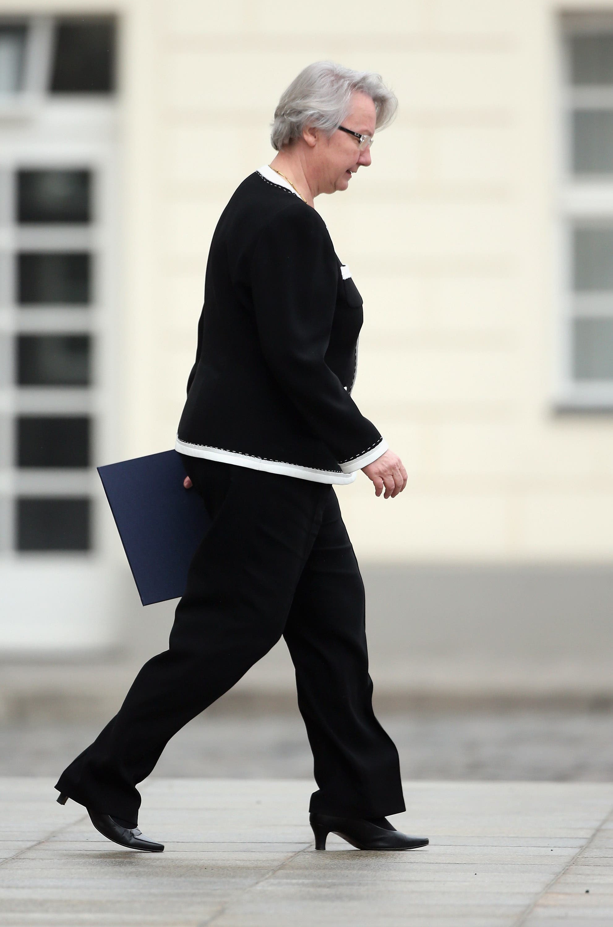 Eine Frau in formeller Kleidung geht draußen auf einem Gehweg. Sie hält eine dunkelblaue Mappe in der Hand. Im Hintergrund ist ein Gebäude mit großen Fenstern zu sehen. Die Szene wirkt professionell und geschäftlich.