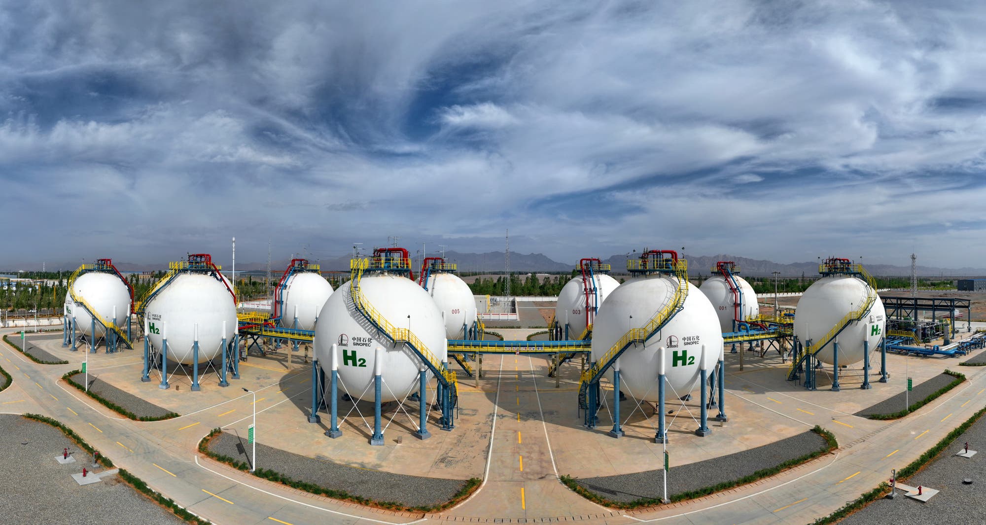 Eine industrielle Anlage mit mehreren großen, kugelförmigen Tanks, die Wasserstoff speichern. Die Tanks sind weiß und mit farbigen Leitern und Plattformen ausgestattet. Im Hintergrund sind Berge und ein bewölkter Himmel zu sehen. Die Umgebung ist sauber und gut organisiert, mit klar markierten Straßen und Gehwegen.