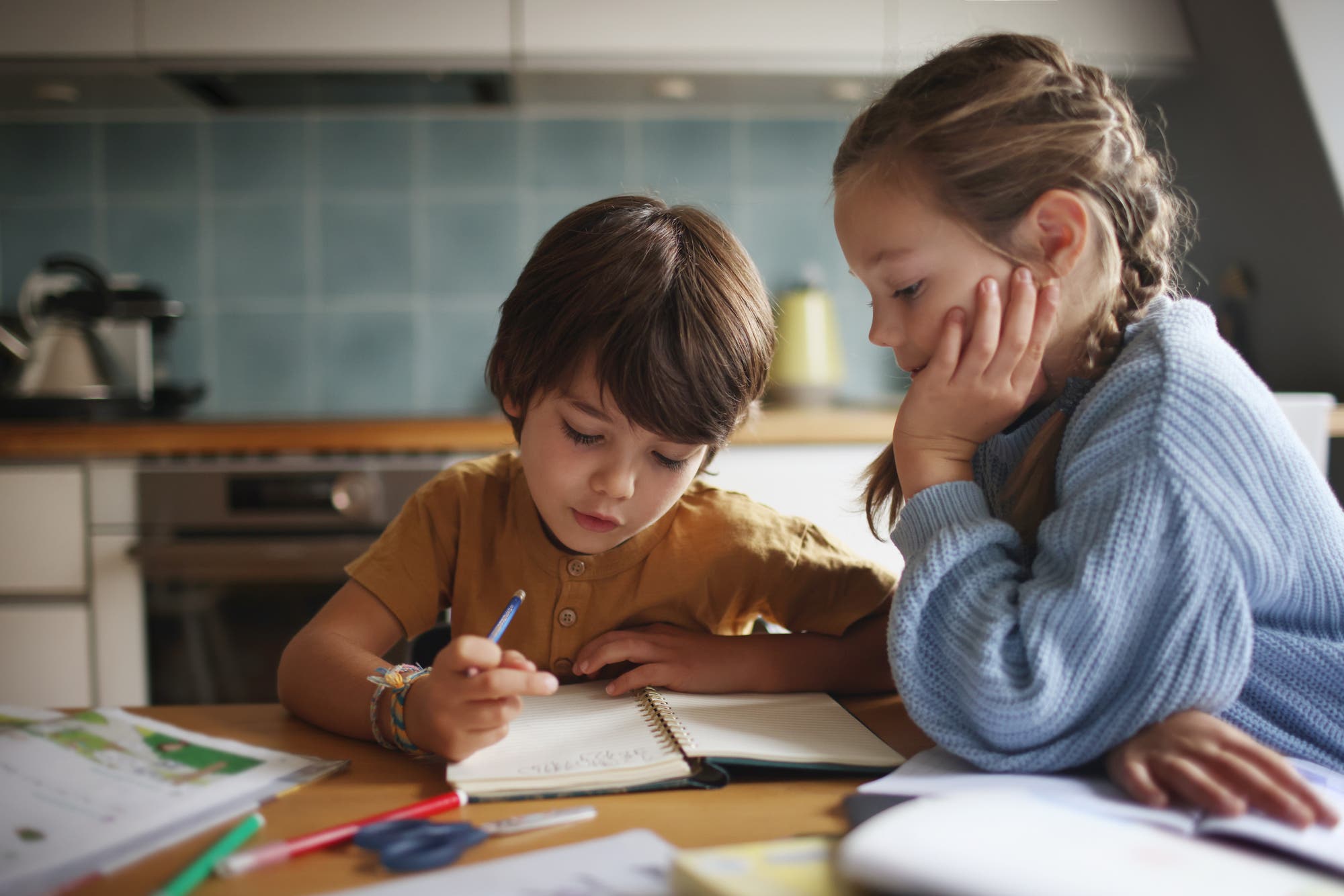 Zwei Kinder sitzen an einem Tisch in einer Küche und arbeiten gemeinsam an einem Notizbuch. Das Kind links schreibt mit einem Stift, während das Kind rechts aufmerksam zuschaut. Auf dem Tisch liegen weitere Schreibutensilien und Papier. Im Hintergrund sind Küchenschränke und eine Arbeitsplatte zu sehen.