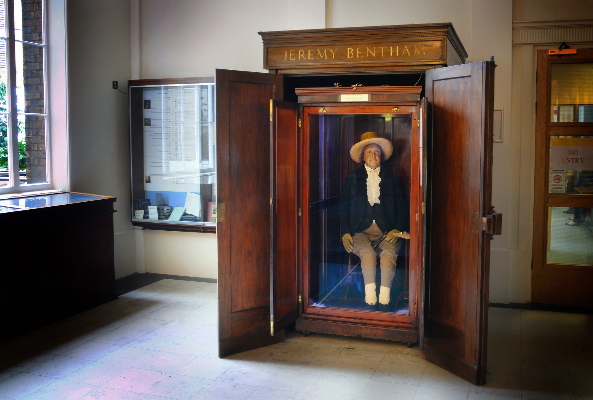 Eine hölzerne Vitrine mit der Aufschrift »Jeremy Bentham« enthält eine bekleidete Figur, die auf einem Stuhl sitzt. Die Figur trägt einen Hut und historische Kleidung. Die Vitrine steht in einem Raum mit weiteren Ausstellungsstücken und einem Fenster, das Tageslicht hereinlässt.