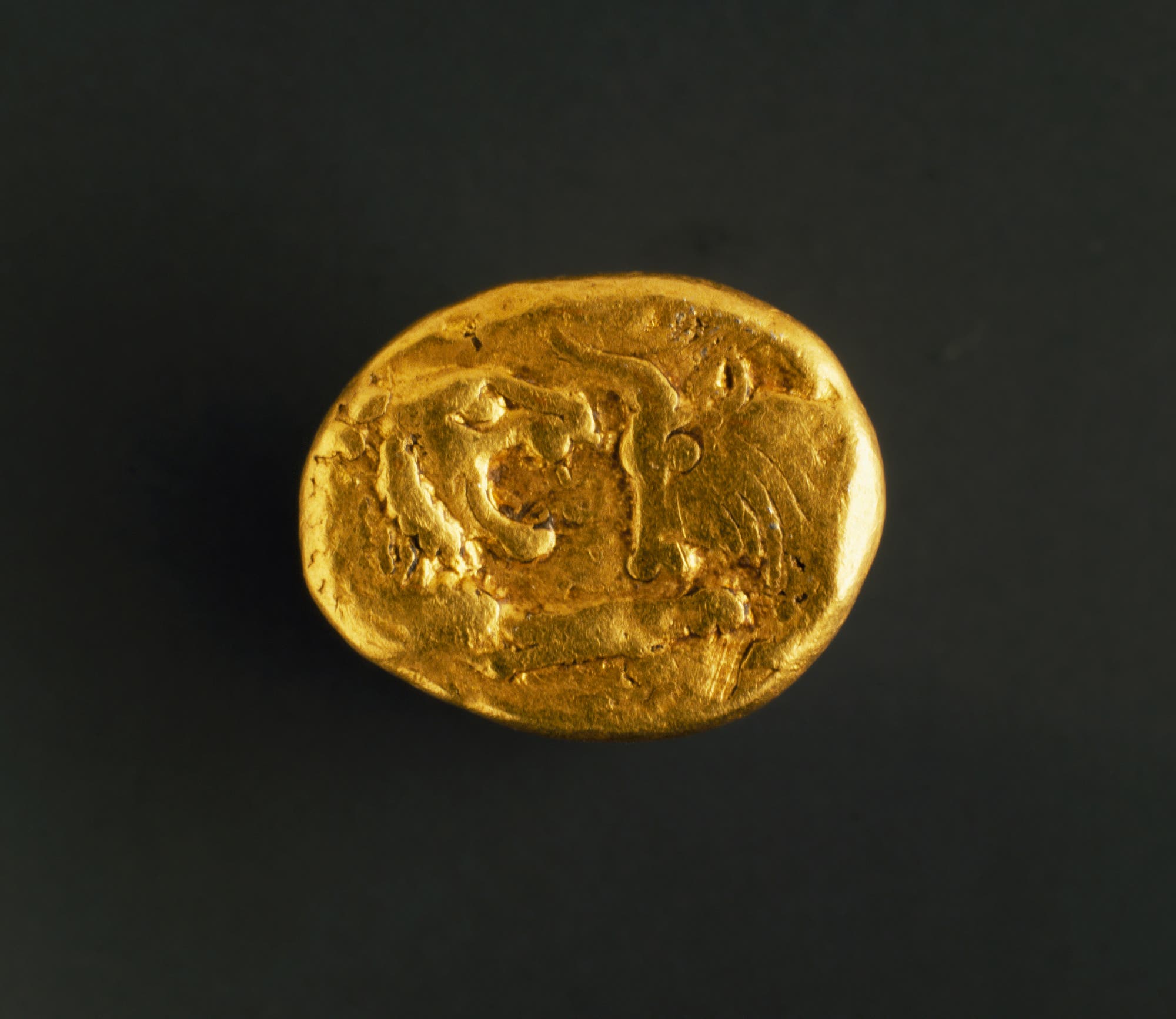 Eine antike Goldmünze mit einem Löwen- und einem Stierkopf als Motiv auf der Vorderseite. Die Münze hat eine unregelmäßige, ovale Form und zeigt Abnutzungsspuren. Der Hintergrund ist dunkel, wodurch die goldene Farbe der Münze hervorgehoben wird.
