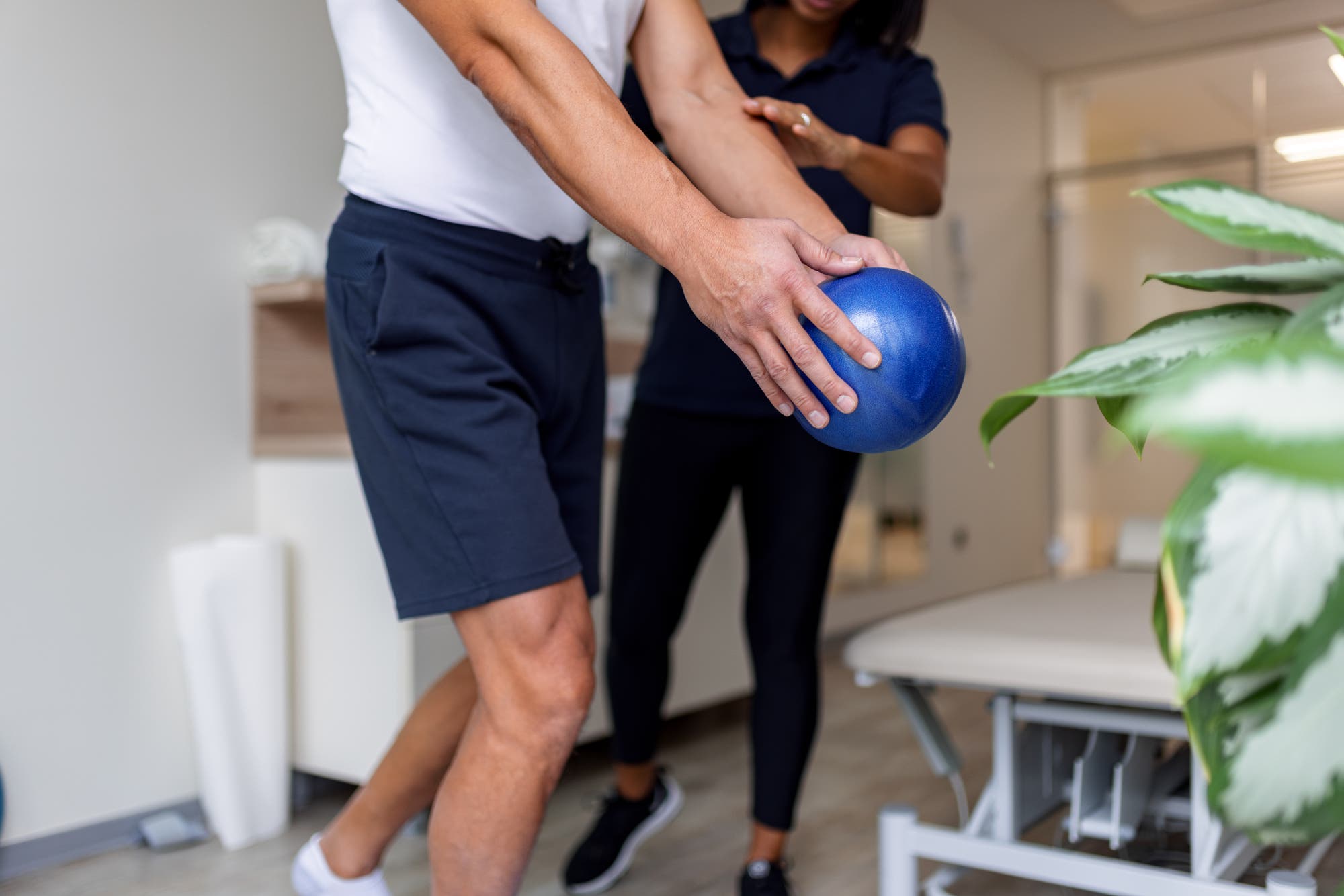Ein Mann in Sportkleidung hält einen blauen Medizinball, während eine Frau in schwarzer Sportkleidung ihm bei einer Übung hilft. Sie befinden sich in einem hellen Raum mit einer Behandlungsliege und einer Pflanze im Vordergrund. Die Szene vermittelt eine physiotherapeutische oder sportliche Trainingseinheit.