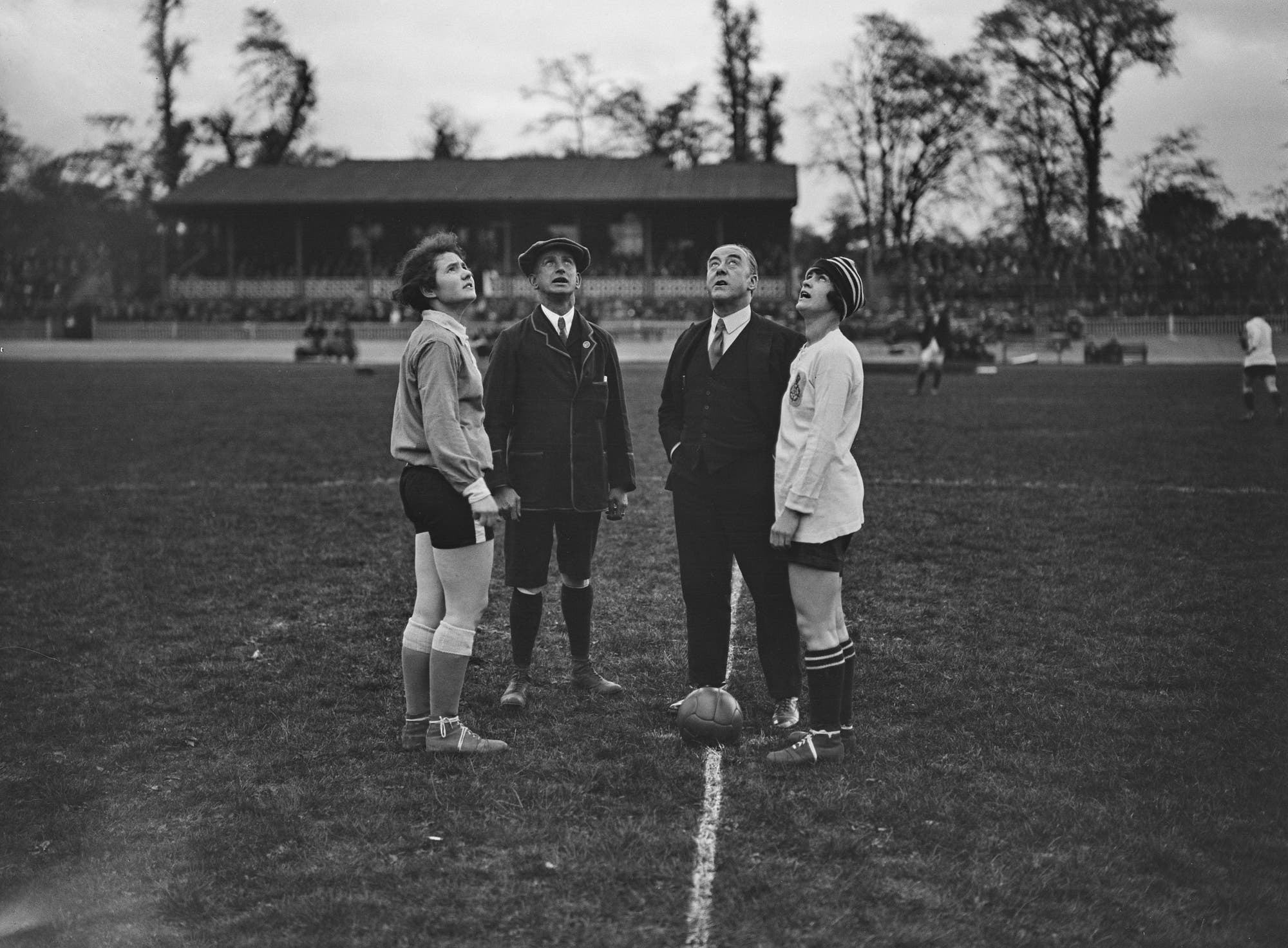 Eine historische Schwarz-Weiß-Aufnahme zeigt zwei Fußballspielerinnen und zwei Offizielle auf einem Spielfeld. Die Spielerinnen stehen sich gegenüber, während die Offiziellen in der Mitte stehen. Alle blicken nach oben, als ob sie einen Münzwurf beobachten. Im Hintergrund sind eine Tribüne und Zuschauer zu sehen. Das Bild vermittelt den Beginn eines Fußballspiels in einer früheren Zeit.