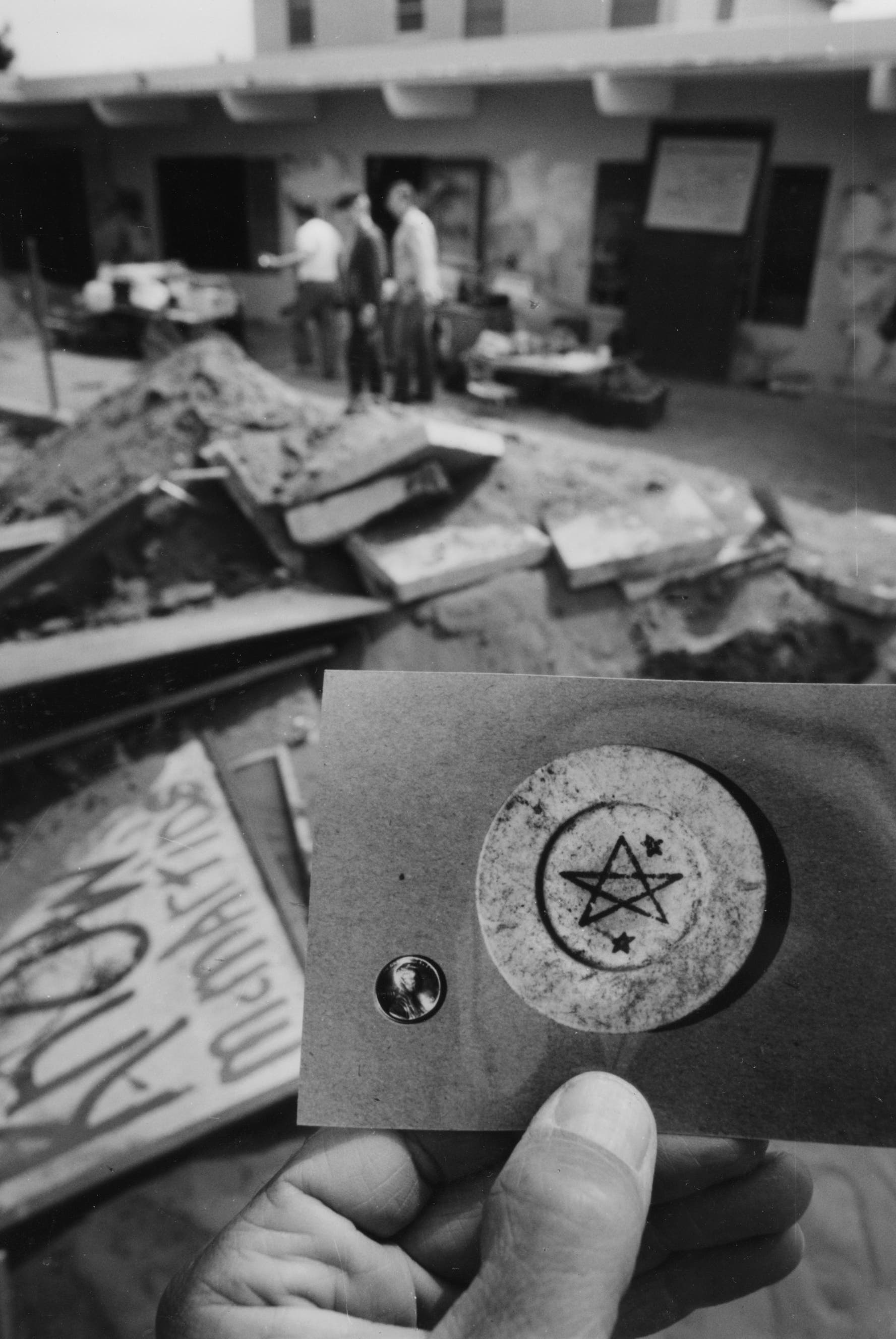 Eine Hand hält ein Foto mit einem kreisförmigen Symbol, das einen Stern und kleinere Sterne zeigt. Im Hintergrund sind unscharf mehrere Personen zu sehen, die vor einem Gebäude stehen. Auf dem Boden liegen Trümmer und Schilder, eines davon mit der Aufschrift "ATOM". Die Szene deutet auf eine Baustelle oder einen Ort der Zerstörung hin.