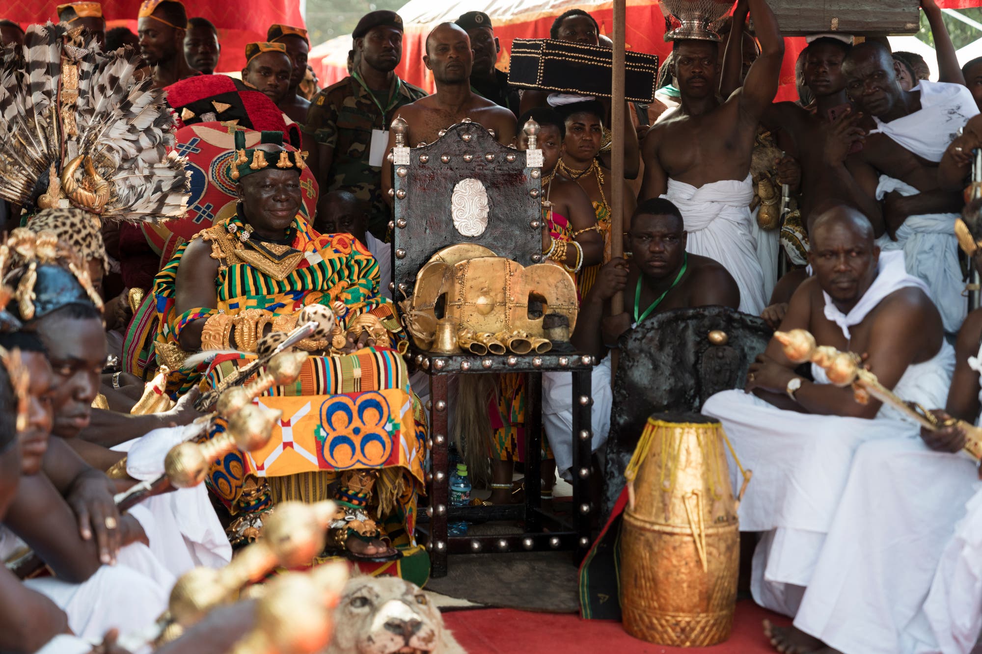 Eine Gruppe von Menschen in traditioneller, farbenfroher Kleidung sitzt und steht um einen reichverzierten Thron. Der Thron ist mit goldenen und dekorativen Elementen geschmückt. Einige Personen halten zeremonielle Gegenstände, und es sind Trommeln und andere kulturelle Artefakte zu sehen. Die Szene findet unter einem roten Baldachin statt, was auf eine festliche oder zeremonielle Veranstaltung hindeutet.