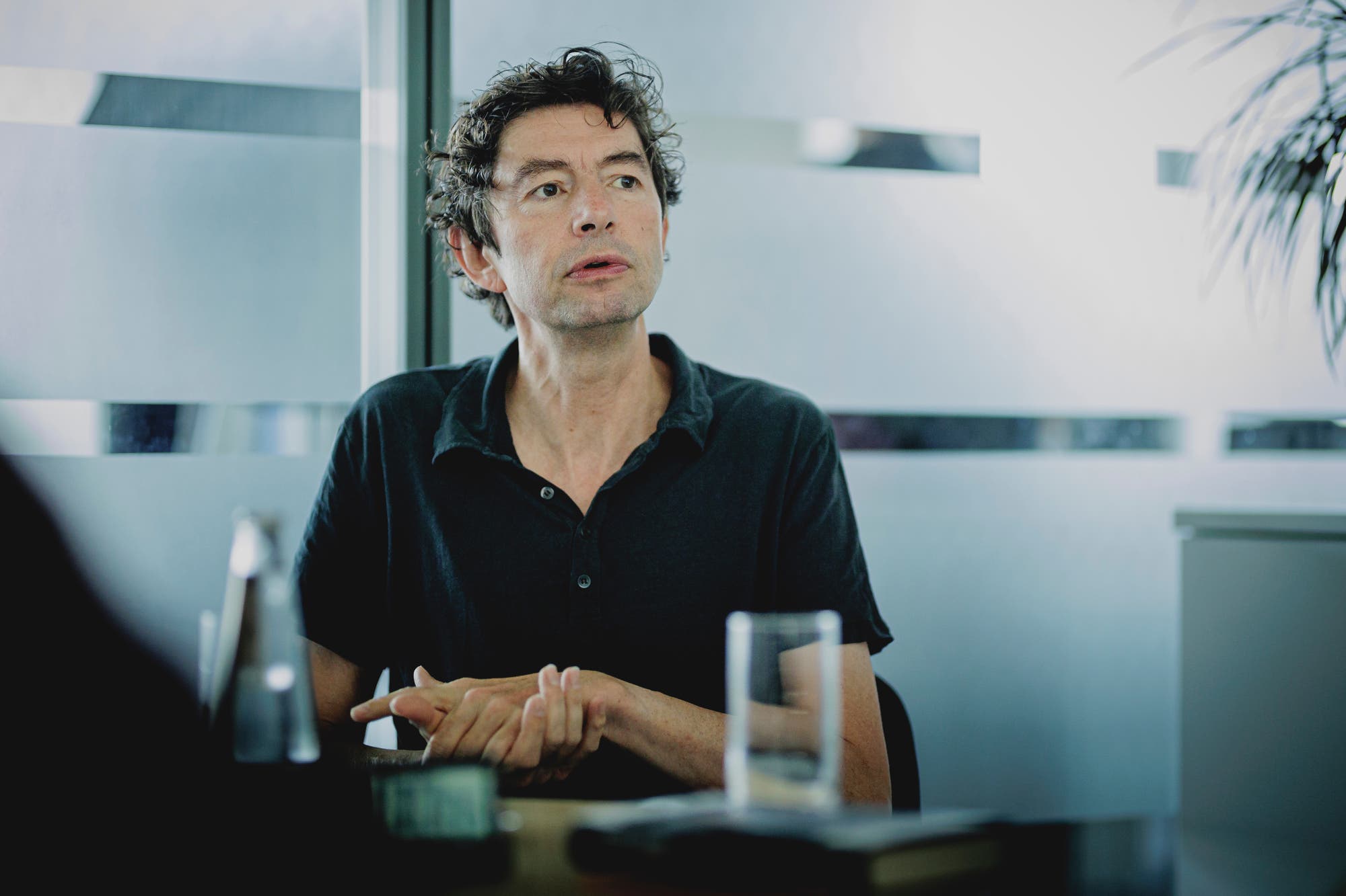 Eine Person sitzt an einem Tisch in einem modernen Büro. Sie trägt ein dunkles Hemd und hat lockiges Haar. Vor der Person stehen ein Glas Wasser und eine Flasche. Im Hintergrund sind Glaswände mit horizontalen Streifen zu sehen. Die Person scheint in ein Gespräch vertieft zu sein.