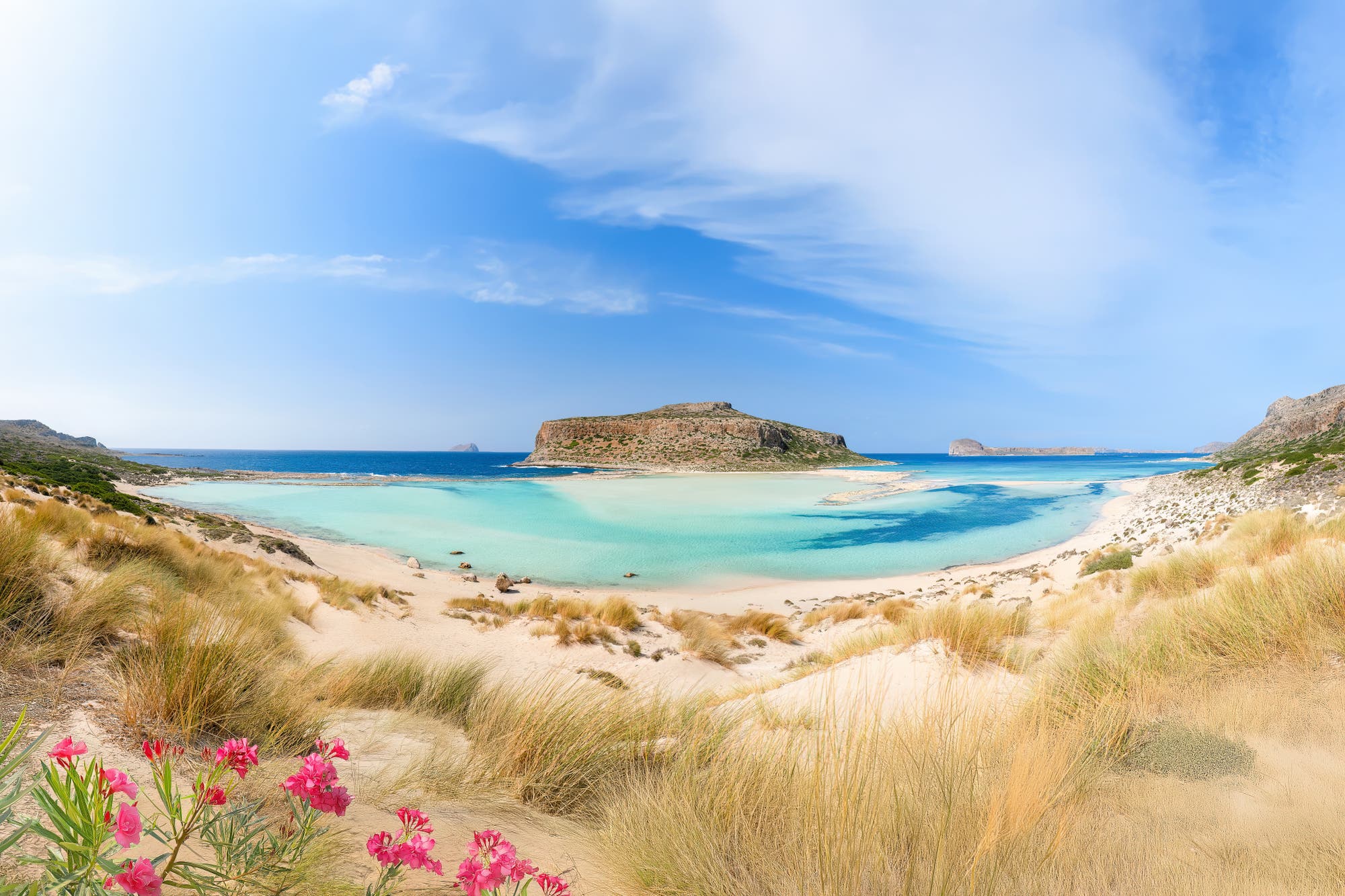 Eine malerische Lagune mit türkisfarbenem Wasser und einem sandigen Strand, umgeben von felsigen Hügeln. Im Vordergrund blühen rosa Blumen zwischen Grasbüscheln. Der Himmel ist klar mit einigen Wolken. Die Szene vermittelt Ruhe und natürliche Schönheit.