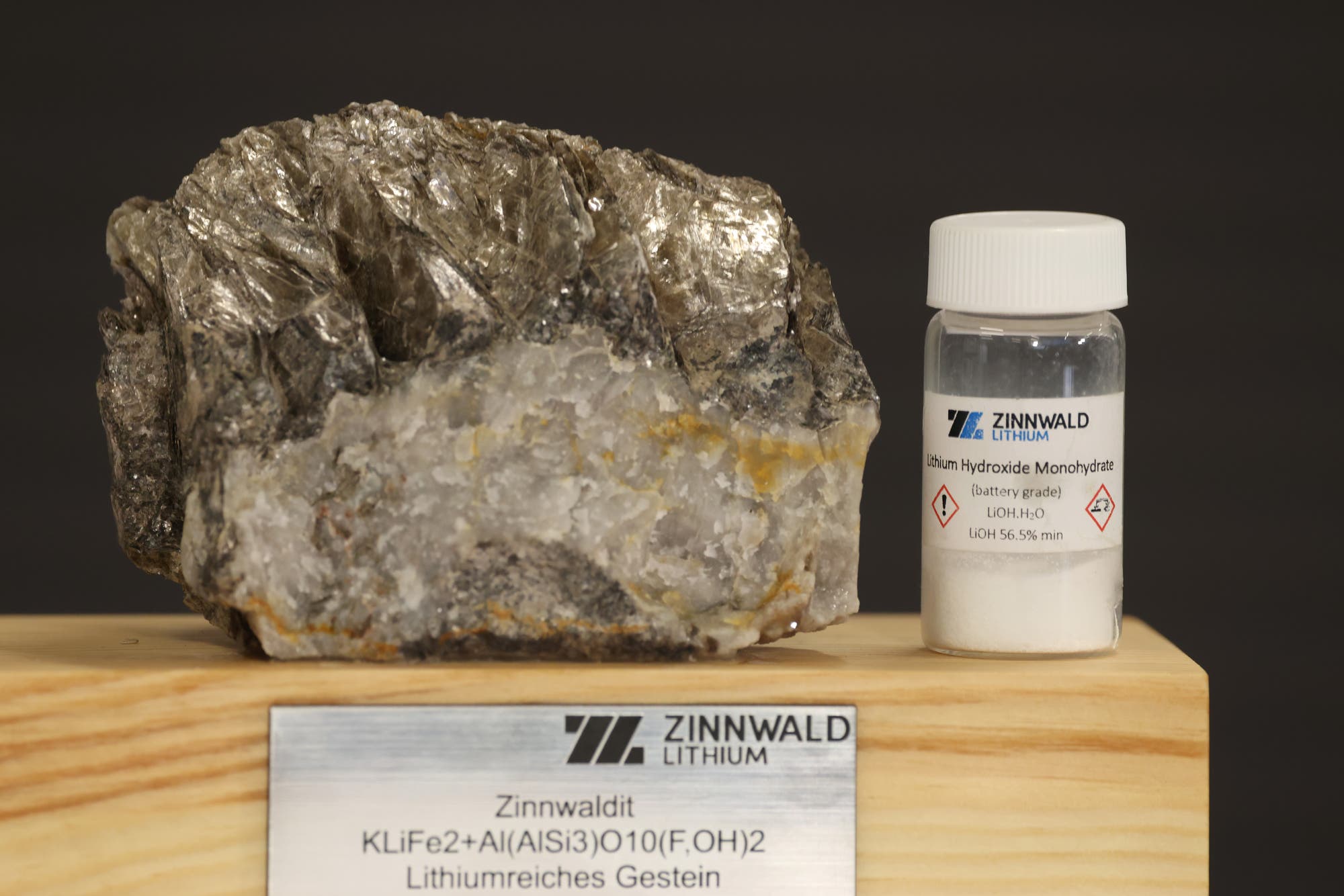 Ein großes Stück Zinnwaldit-Gestein und ein kleines Fläschchen mit Lithiumhydroxid-Monohydrat stehen nebeneinander auf einem Holzsockel. Das Etikett auf dem Fläschchen zeigt die Aufschrift "Zinnwald Lithium" und "Lithium Hydroxide Monohydrate (battery grade) LiOH.H2O LiOH 56.5% min". Die Plakette auf dem Sockel beschreibt das Gestein als "Zinnwaldit KLiFe2+Al(AlSi3)O10(F,OH)2 Lithiumreiches Gestein".