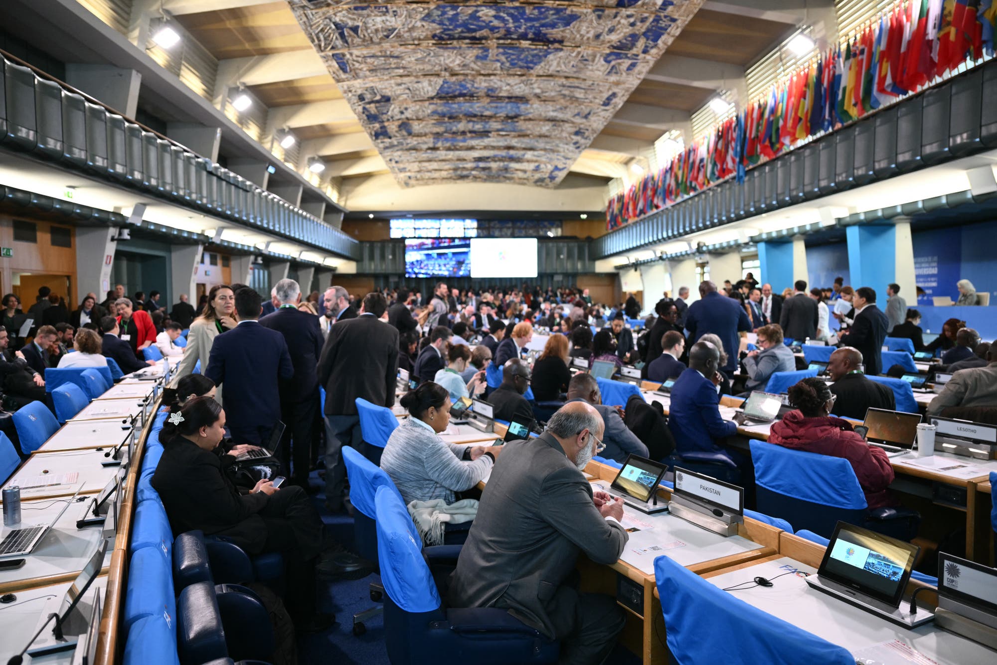 Eine große Konferenzhalle voller Menschen, die an einem internationalen Treffen teilnehmen. Die Teilnehmer sitzen an Tischen mit Laptops und Namensschildern. An den Wänden hängen zahlreiche Flaggen verschiedener Länder. Im Hintergrund sind Bildschirme mit Präsentationen zu sehen. Die Decke ist mit einem kunstvollen blauen Muster dekoriert. Die Atmosphäre ist geschäftig und konzentriert.
