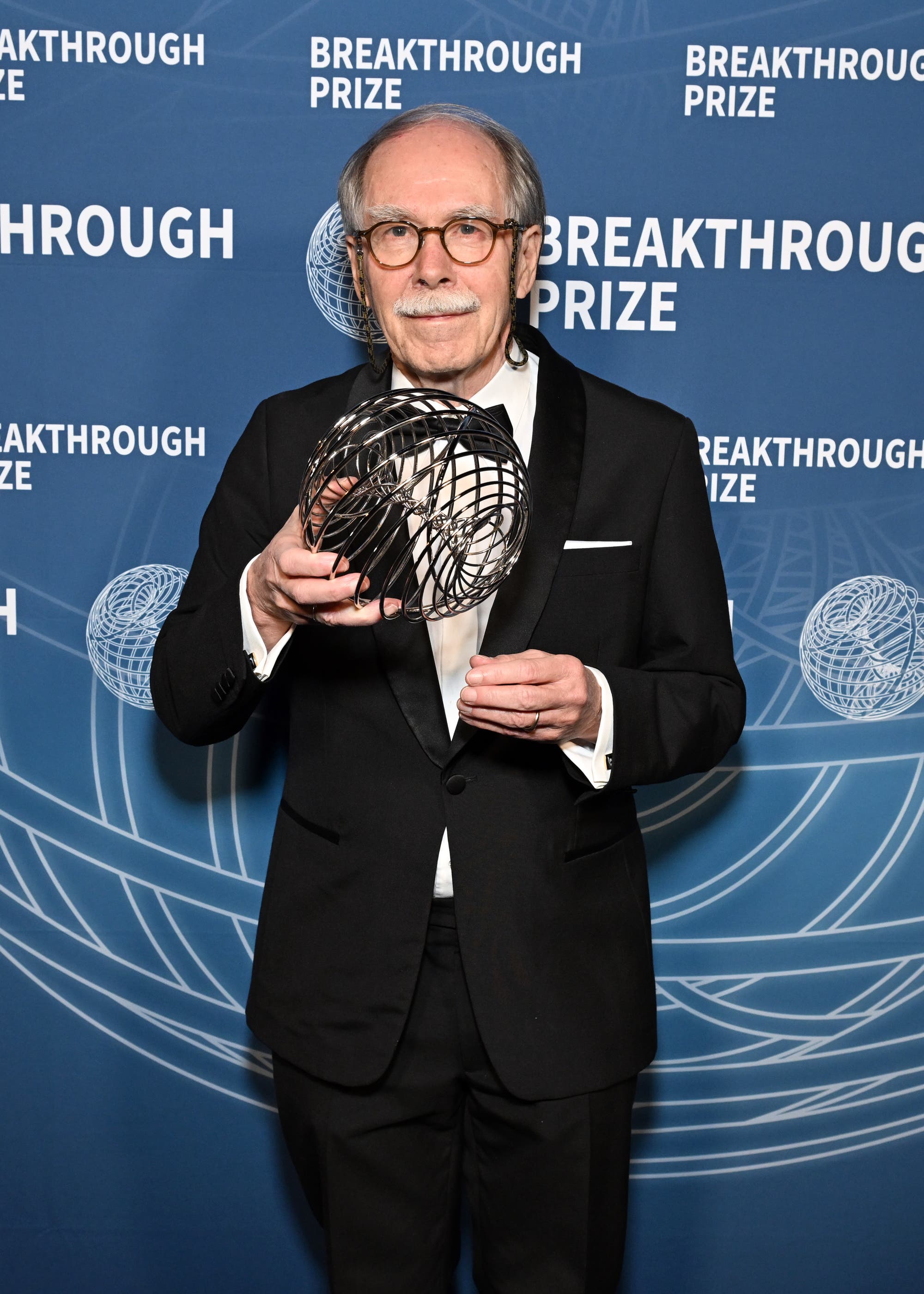 Gerard 't Hooft, ein älterer Mann mit Schnurrbart in einem schwarzen Anzug, hält eine Trophäe mit einem ein komplexen, kugelförmigen Design, den Breakthrough Prize