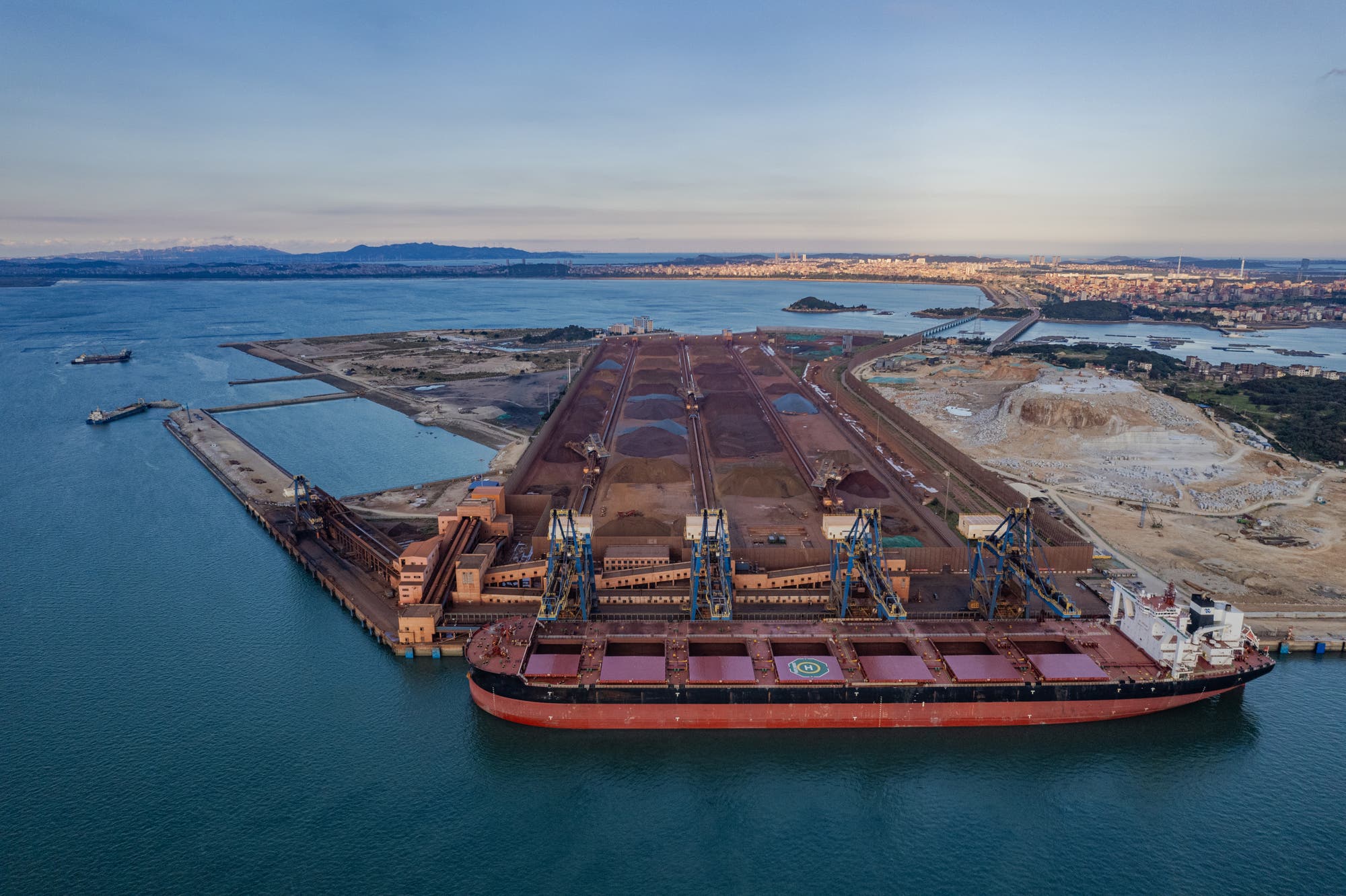 Luftaufnahme eines großen Frachtschiffs, das an einem Industriehafen anlegt. Im Hintergrund sind mehrere Lagerplätze mit Rohstoffen zu sehen, die von Kränen bearbeitet werden. Die Szene zeigt eine Küstenlinie mit Wasser, Land und einer Stadt im Hintergrund. Der Himmel ist klar, und die Umgebung wirkt ruhig.