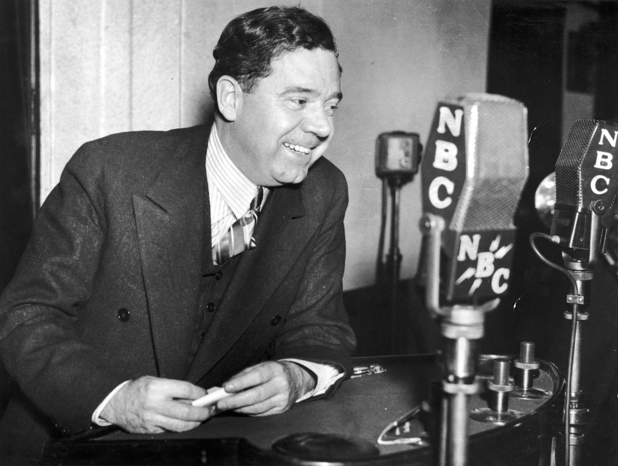 Ein Mann in einem Anzug – Huey Long – steht lächelnd vor mehreren Mikrofonen mit dem NBC-Logo. Er hält einen kleinen Gegenstand in den Händen und scheint in einer Radiosendung zu sprechen. 